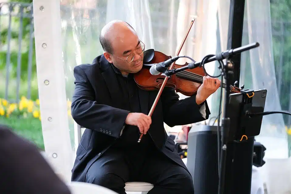 Man in black suit playing violin in front of a microphone with a music stand and outdoor background.