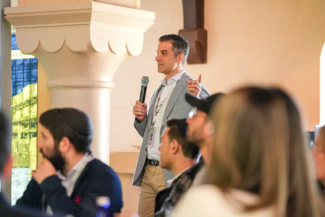 Man in gray blazer holding a microphone and speaking to an audience seated indoors.