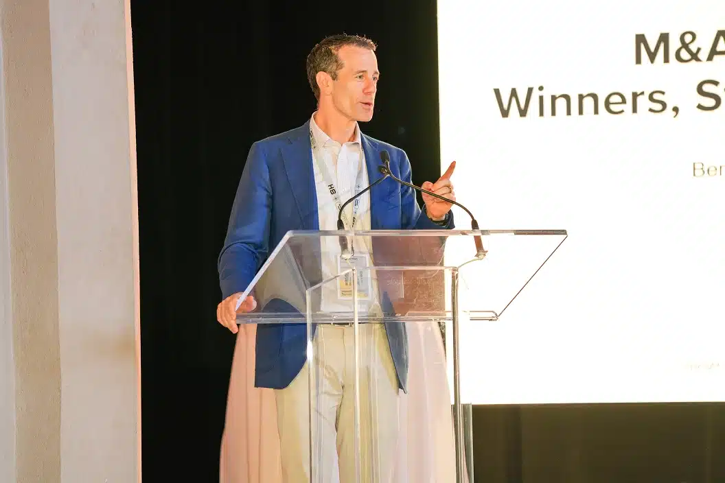 Man in blue blazer speaking at a clear podium beside a large screen displaying text.