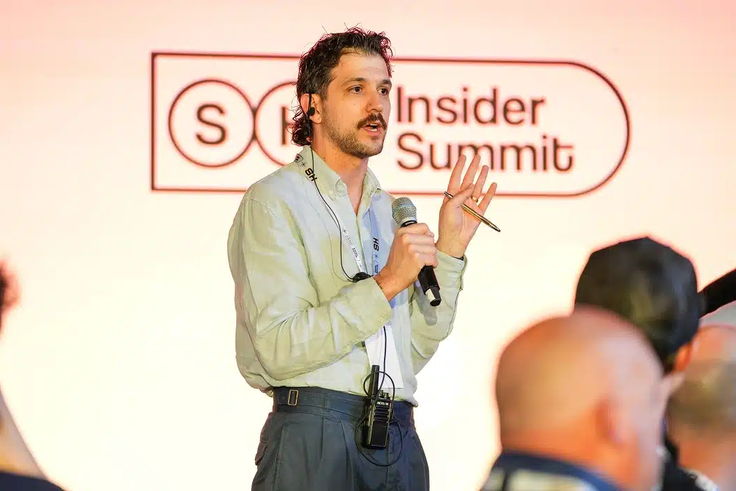 Man holding a microphone and pen speaking at an event with a blurred audience in the foreground and a backdrop reading 'Insider Summit.'