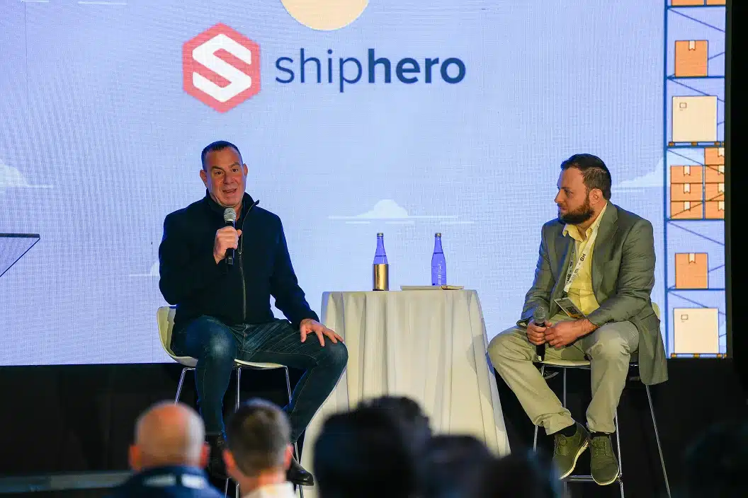 Two men seated on stage during a discussion event with a ShipHero logo displayed on the screen behind them.