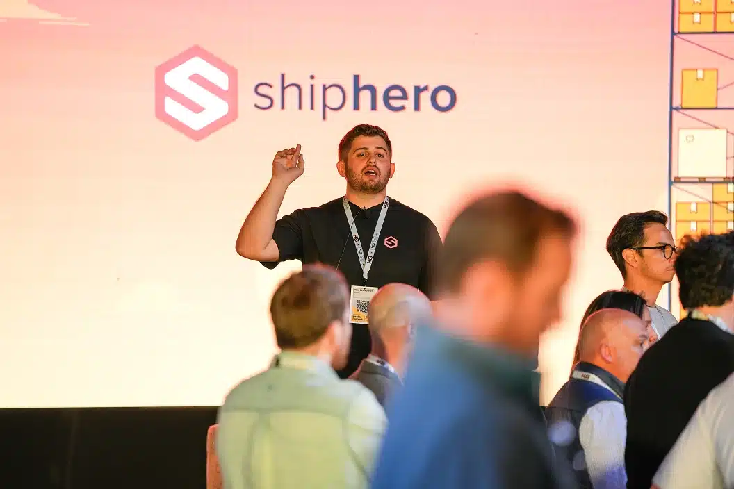 Man in a black shirt speaking on stage with ShipHero logo projected behind him at a conference.