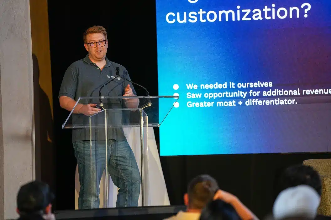 Man wearing glasses and a gray polo shirt speaking at a podium with a presentation slide displaying bullet points about customization in the background.