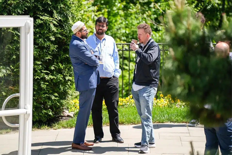Three men engaged in conversation outdoors on a sunny day, with one man drinking from a cup.