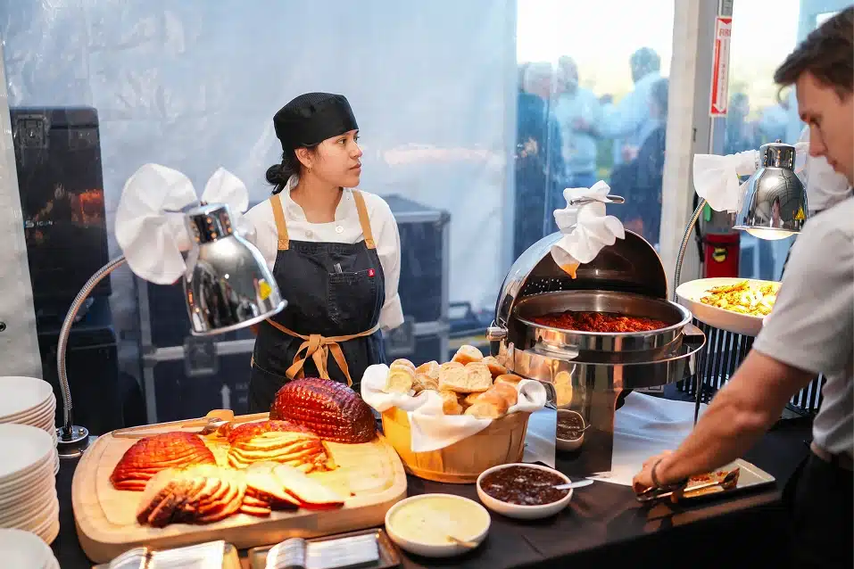Woman chef in black hat and apron stands behind buffet table with sliced ham, bread rolls, and hot dishes while a man serves food.