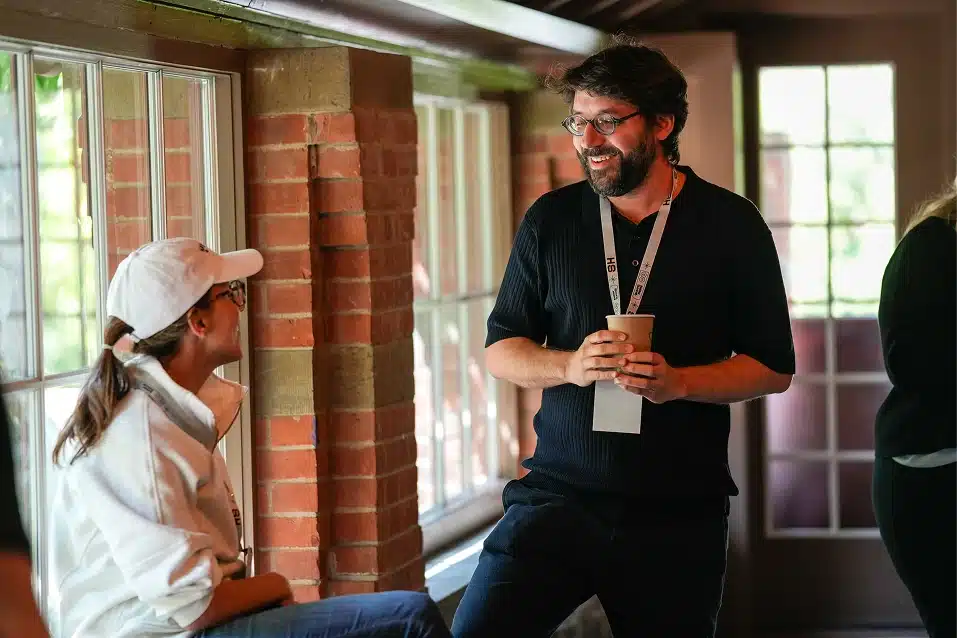 Two people having a friendly conversation indoors, one sitting near a window and the other standing holding a cup.