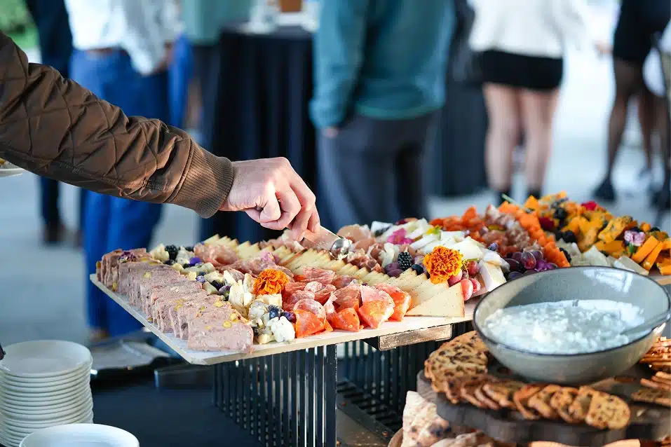 Person in brown jacket picking food from a charcuterie board with cheese, meats, fruits, and edible flowers at an event.