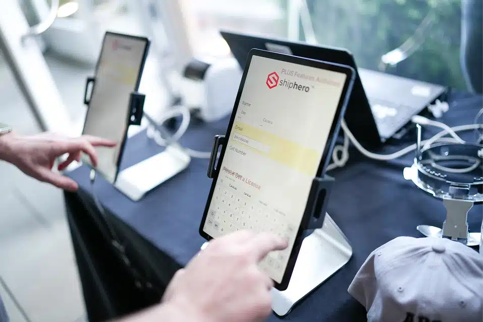 Person interacting with a touchscreen tablet displaying the ShipHero app interface on a black table with another device and laptop in the background.