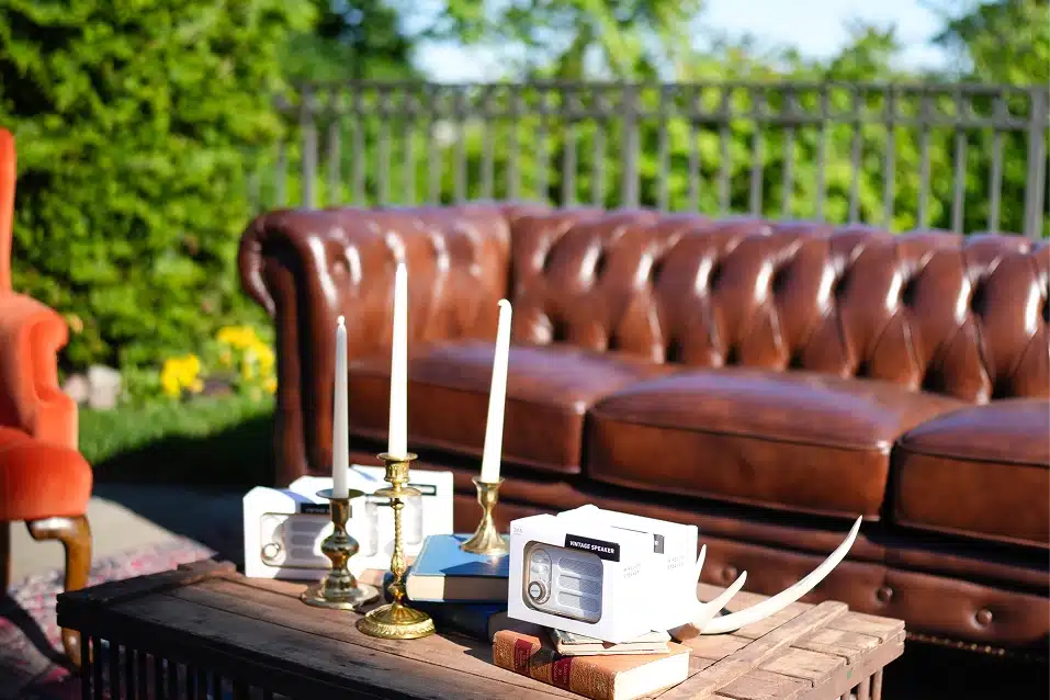 Outdoor seating area with a brown leather tufted sofa, wooden table holding candles, books, vintage speakers, and antlers.