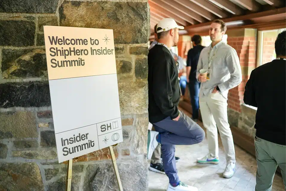 Sign reading 'Welcome to ShipHero Insider Summit' next to a stone wall, with people standing and conversing in a hallway.