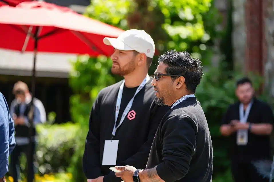 Two men with conference badges standing outdoors near a red umbrella and greenery.