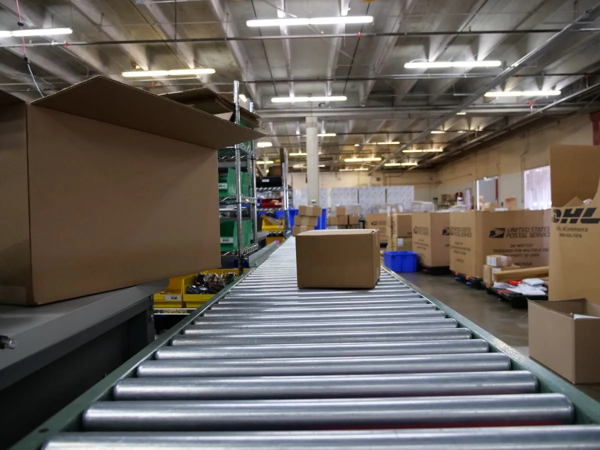 Cardboard box on a roller conveyor belt in a warehouse filled with shipping boxes and shelves.