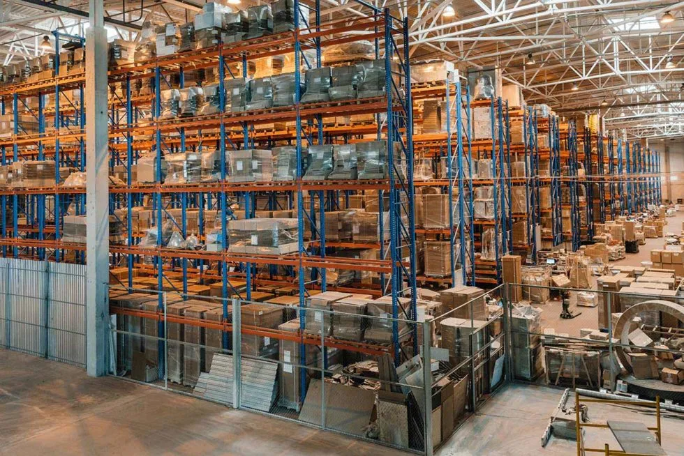 Large warehouse with tall shelves stacked with pallets and boxes under bright industrial lights.