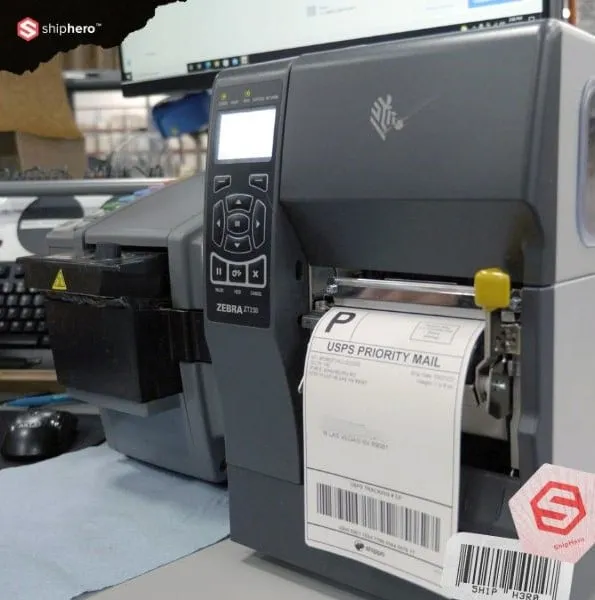 Zebra printer printing a USPS Priority Mail label in an office setting with computer screens in the background.