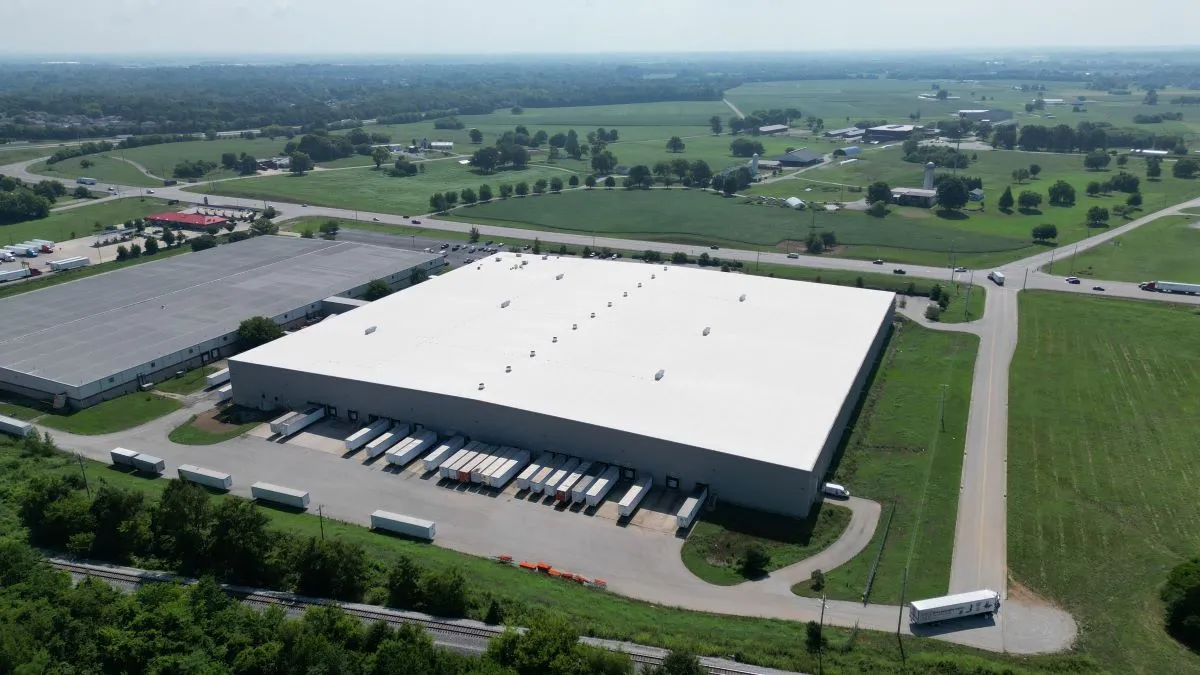 Aerial view of a large warehouse with several trucks parked at loading docks and surrounding green fields.