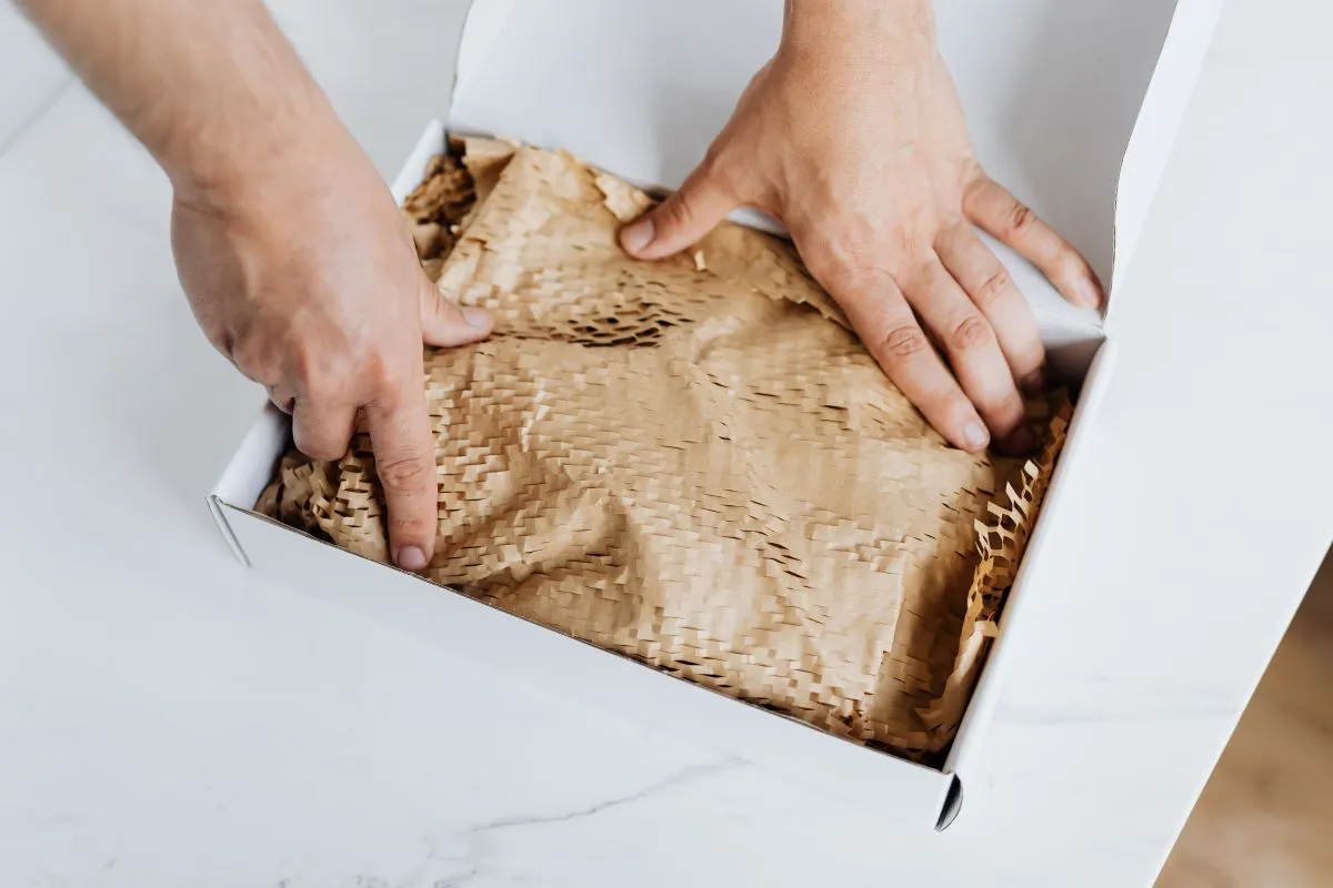 Two hands packing or unpacking a white box lined with brown textured protective paper.