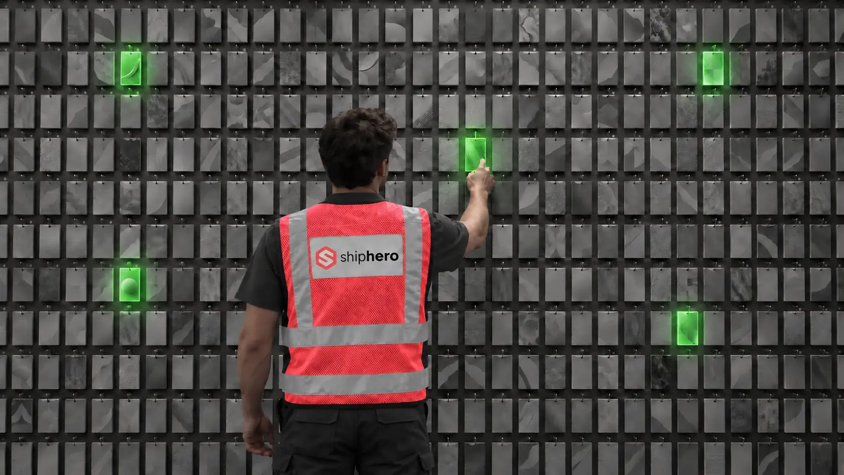 Worker in a red ShipHero safety vest, seen from behind, pointing at a glowing green card on a wall of gray cards