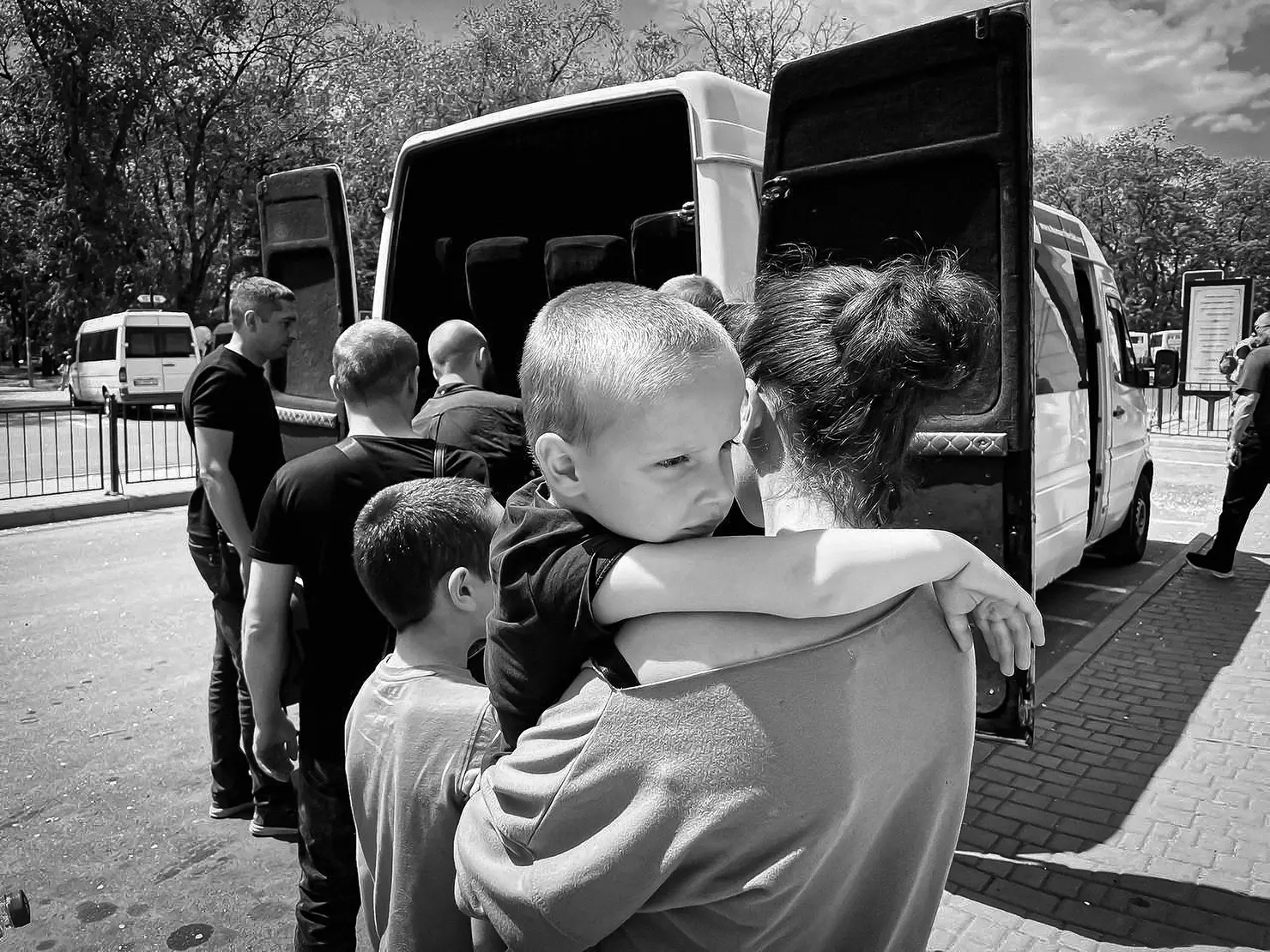 Schwarz-weiss Foto einer wartenden Gruppe von Menschen, darunter eine Frau, die ein Kind trägt, vor einem offenen Minibus.