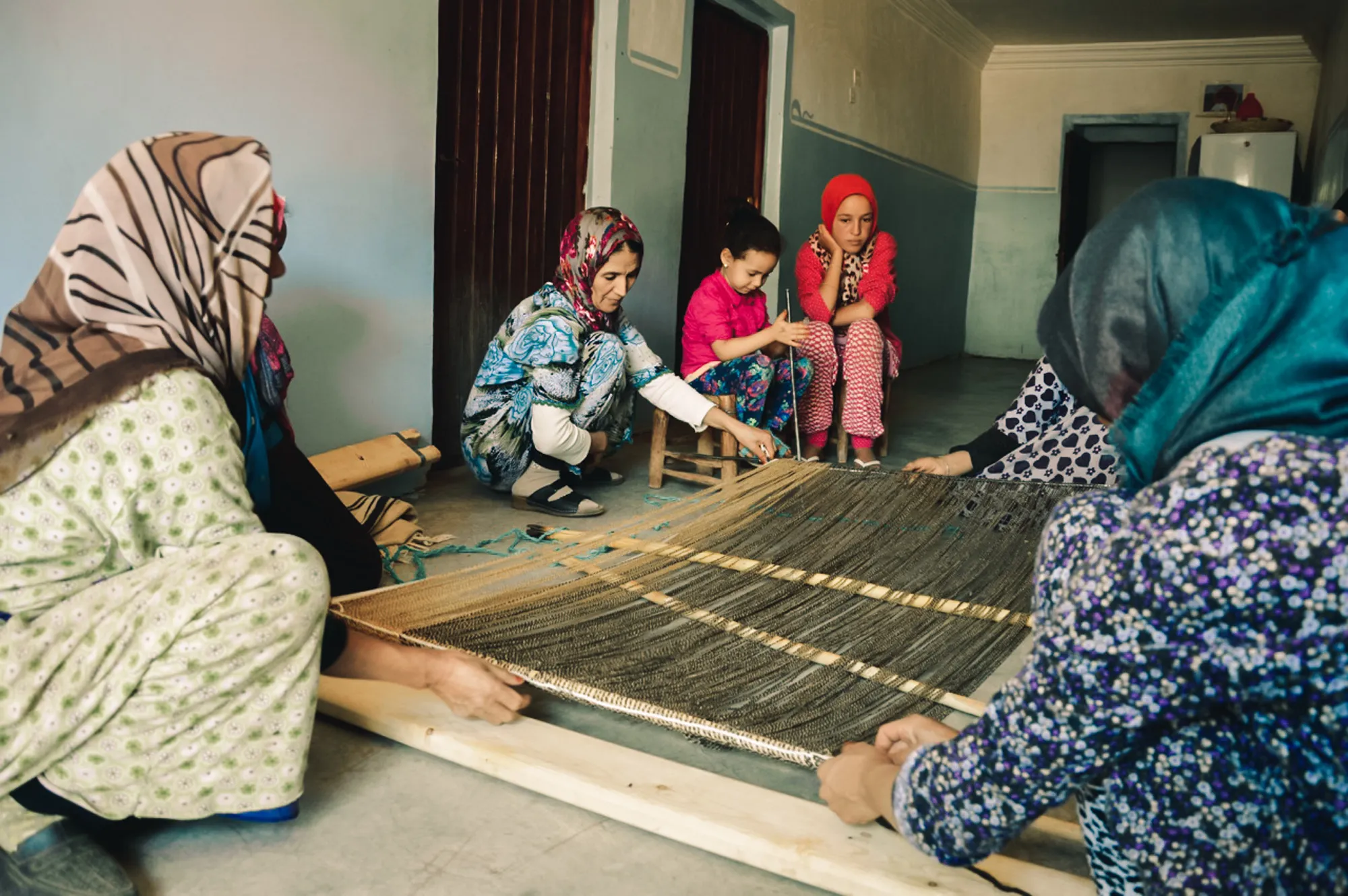 Groupe de femmes amazighes marocaines et une jeune fille réunies autour d’un métier à tisser horizontal au sol, confectionnant un tapis traditionnel marocain dans une pièce aux murs bleus, dans la vallée des Aït Bougmez, Haut Atlas – Shop Touda.