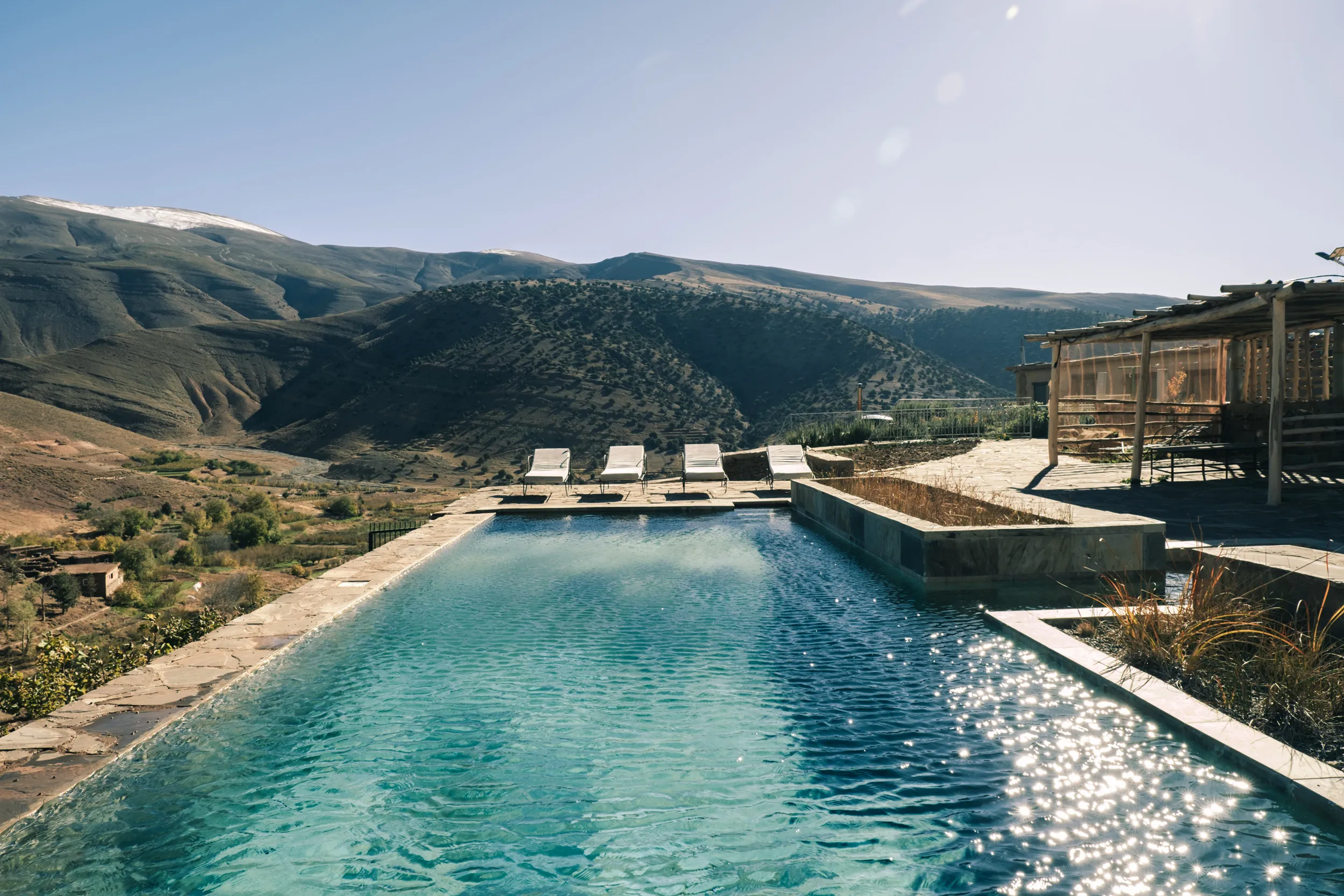 Piscine de touda ecolodge avec chaises allongées au bord et montagnes de haut atlas centrale en arrière-plan dans la vallée des ait Bougemez, region d'Azilal au Maroc.