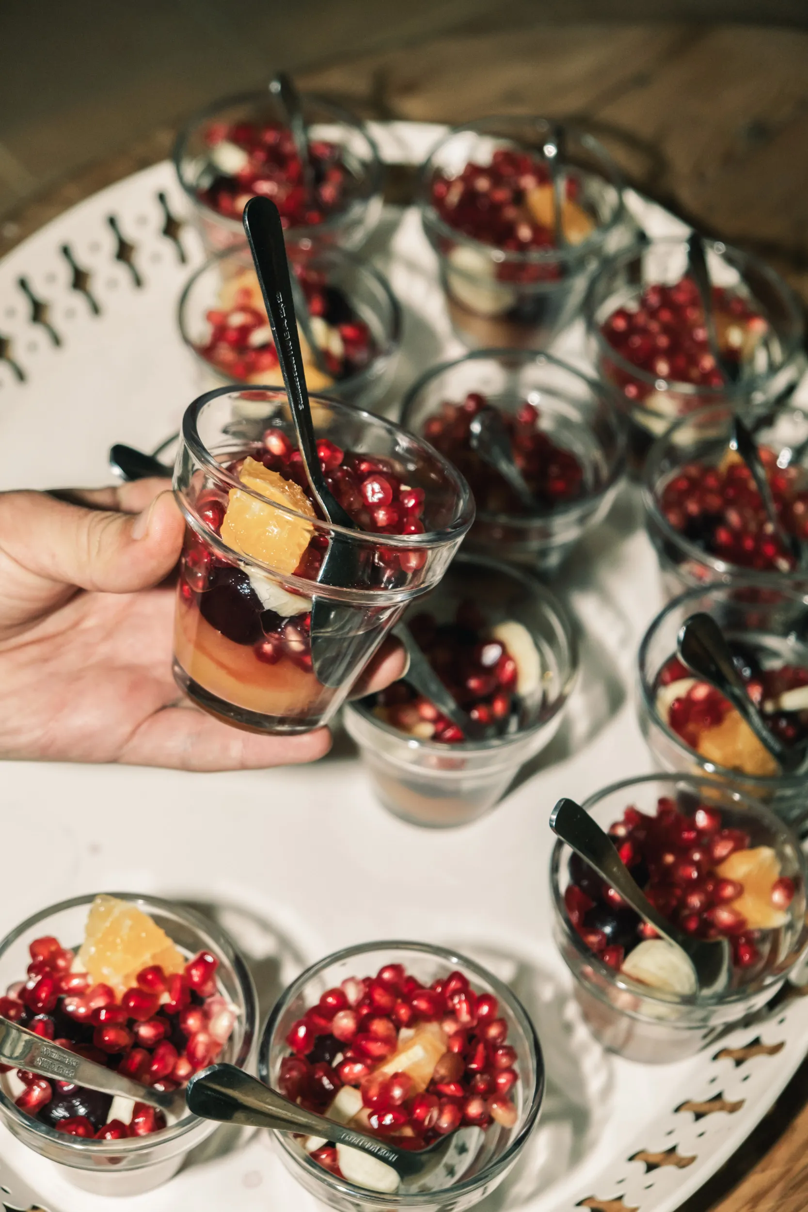 Cuisinière de Touda ecolodge tenant un petit verre transparent rempli de fruits frais et bio du potager, notamment des grains de grenade et des morceaux d'orange, avec plusieurs verres similaires sur un plateau pour une famille en séjour au haut atlas.
