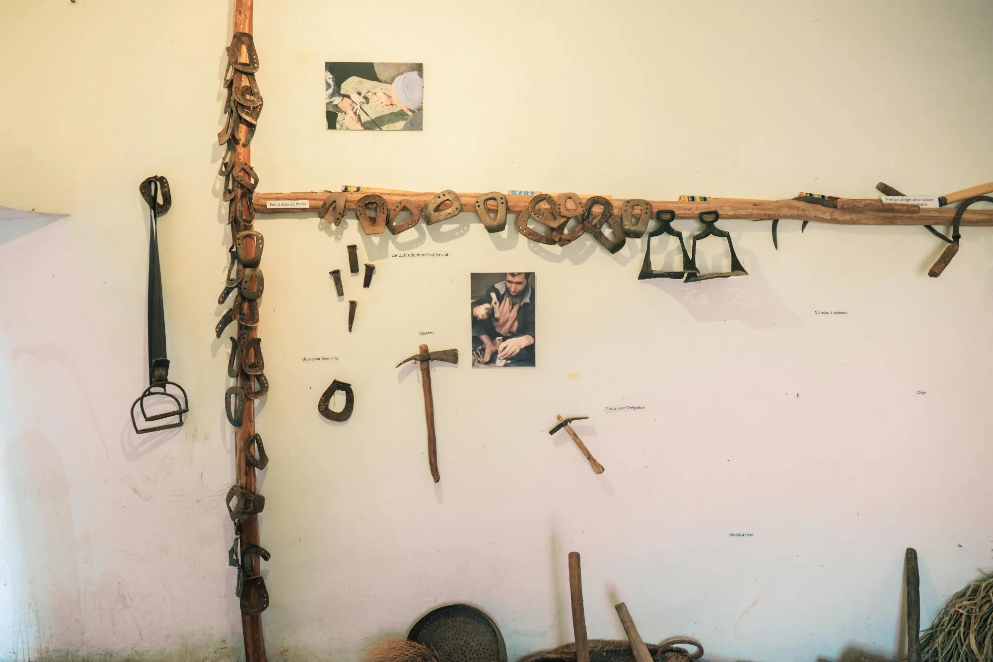 Mur de l'ecomusée de la vallée des ait Bougemez affichant divers outils traditionnels de maréchal-ferrant et d'agriculture amazigh, y compris des fers à ânes, des marteaux et des clous, avec des photos illustratives.