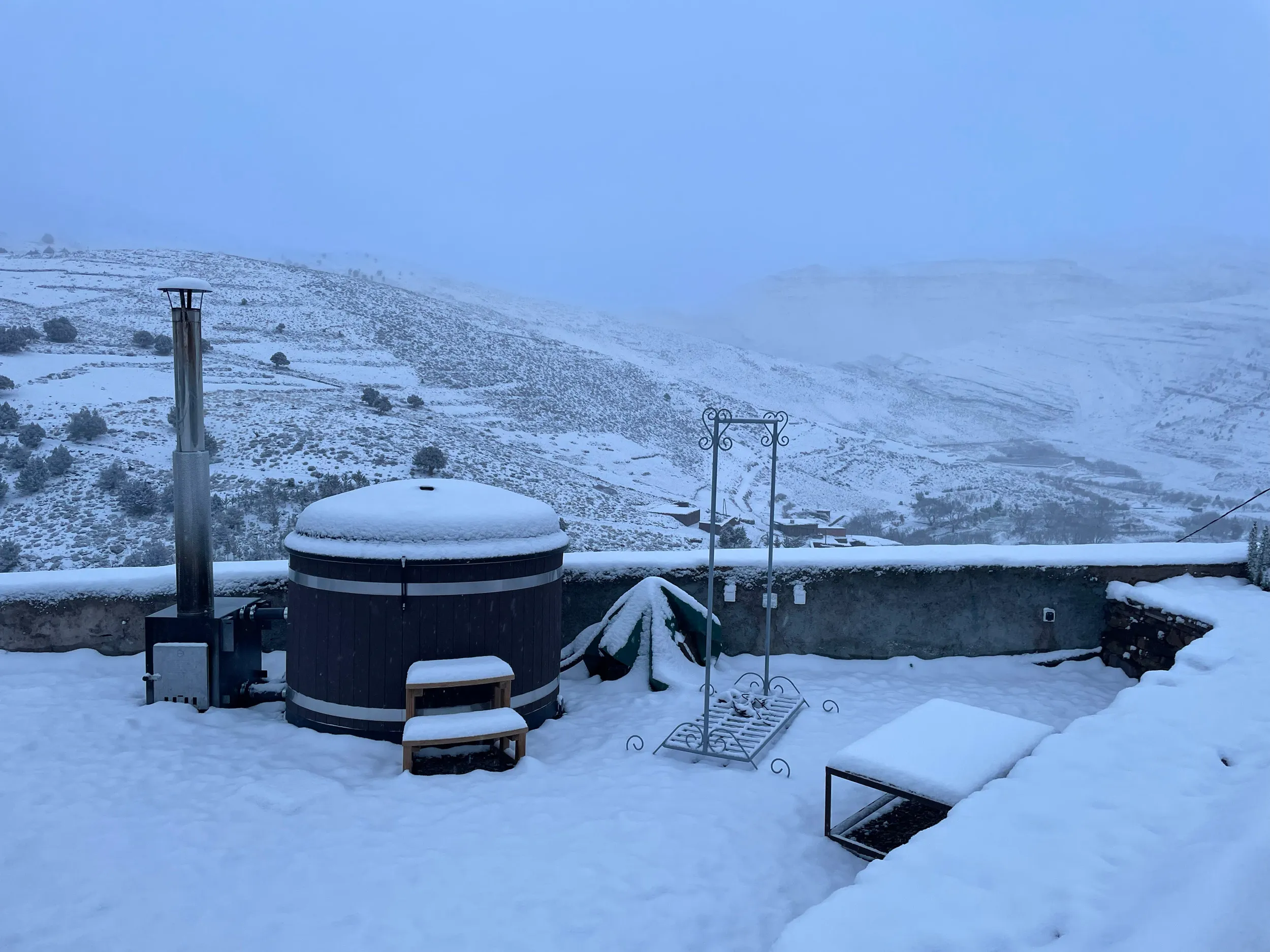 Terrasse de touda ecolodge enneigée avec le seul bain nordique au maroc, des bancs couverts de neige, et une vue sur des collines de la vallée heureuse de bougmaz à l'atlas centrale du Maroc.