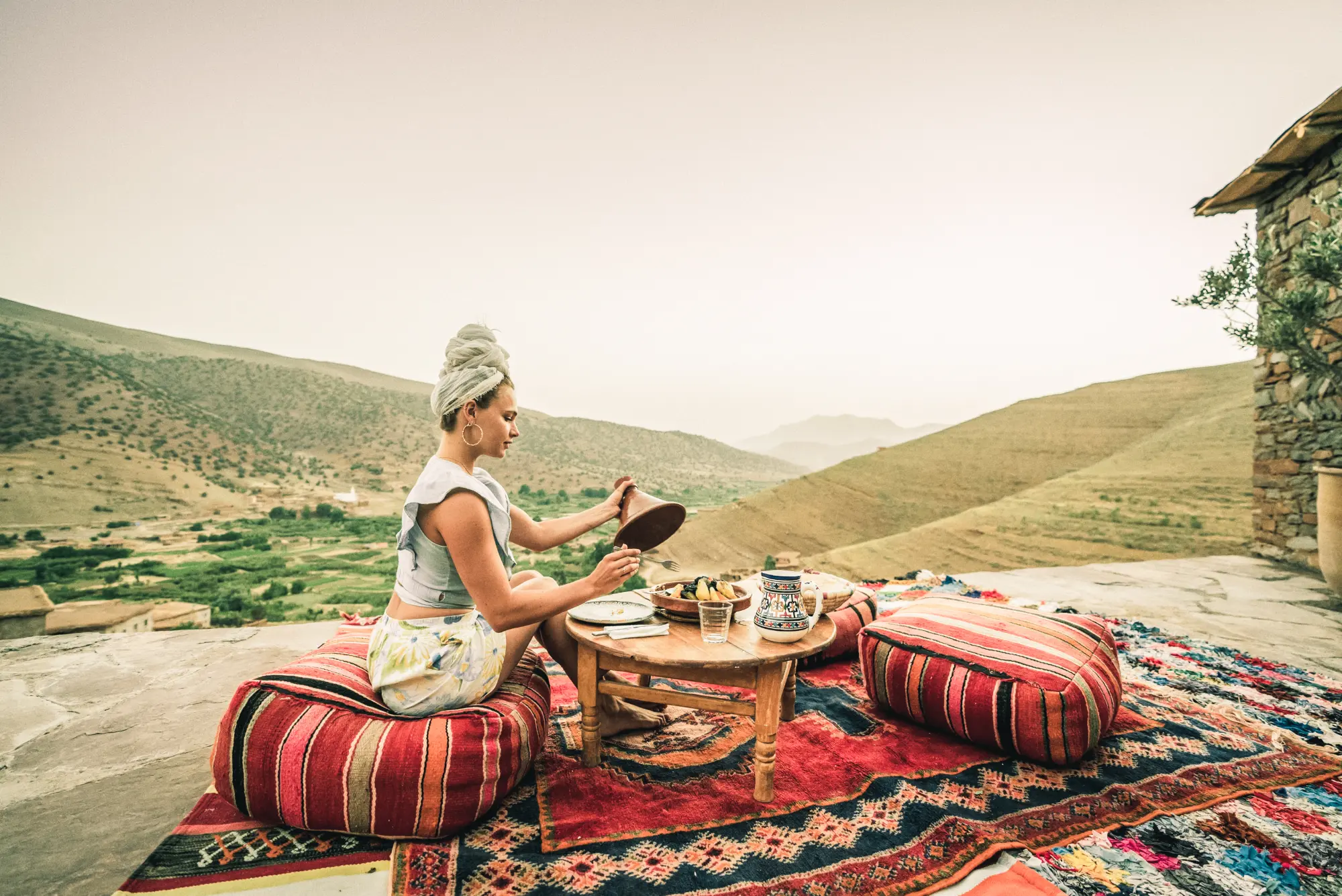 Une jeune femme assise sur des poufs traditionnels déguste un tajine sur une terrasse en pierre, offrant une vue panoramique imprenable sur la vallée heureuse et les montagnes du Haut Atlas.