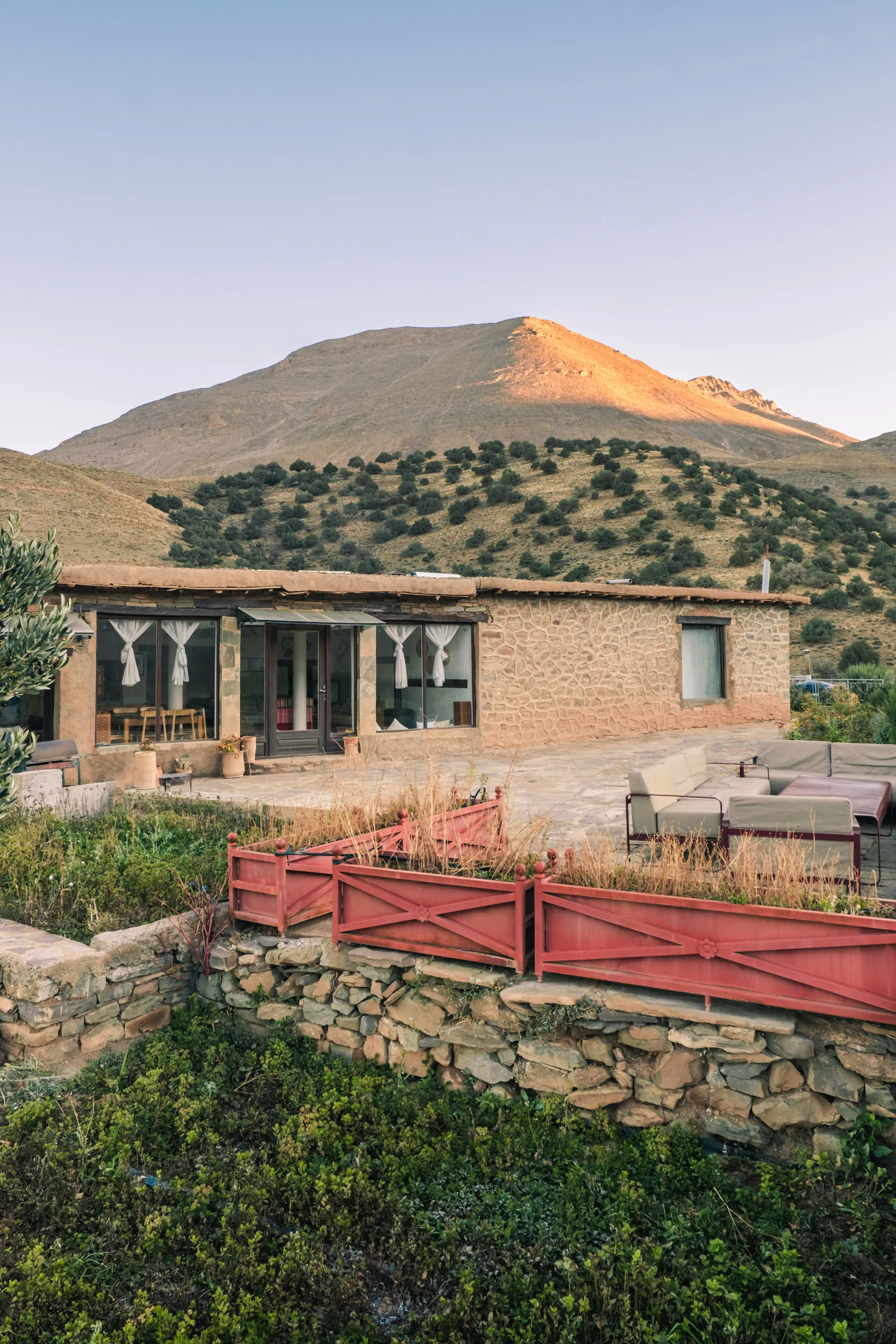 Touda ecolodge un auberge au haut atlas marocain dans la vallée des ait bougemez construit en pierre avec une terrasse meublée et une barrière rouge, au pied d'une colline verte sous un ciel clair.