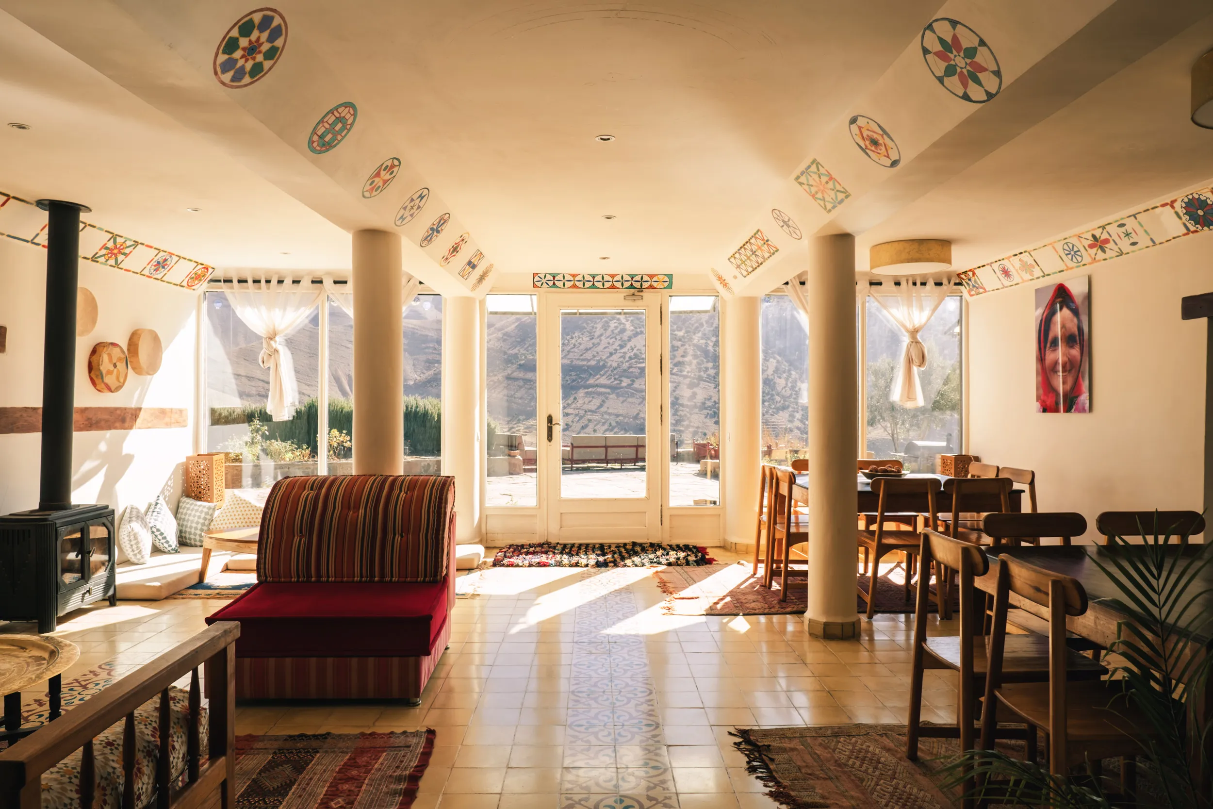 Intérieur de Touda ecolodge avec un salon canapés, table à manger en bois, poêle à bois et grandes fenêtres donnant sur un le village de Zawiat Oulmzi dans la vallée Bougemez.