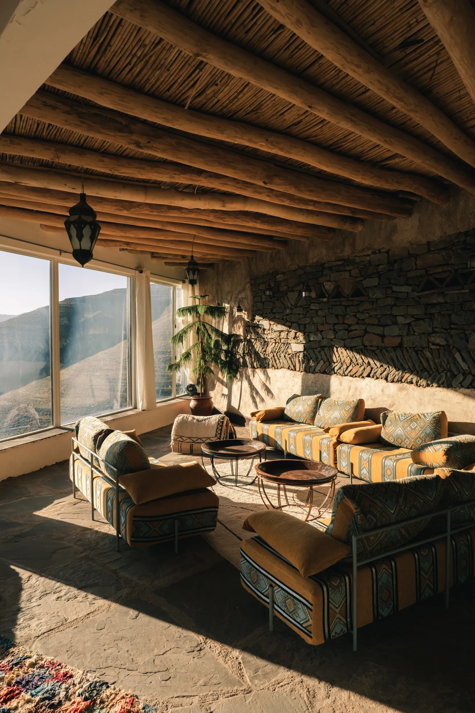 Salon rustique avec canapés colorés, murs en pierre, plafond en poutres de bois, grandes fenêtres offrant une vue sur la vallée ait Bougemez dans le haut atlas Marocain.