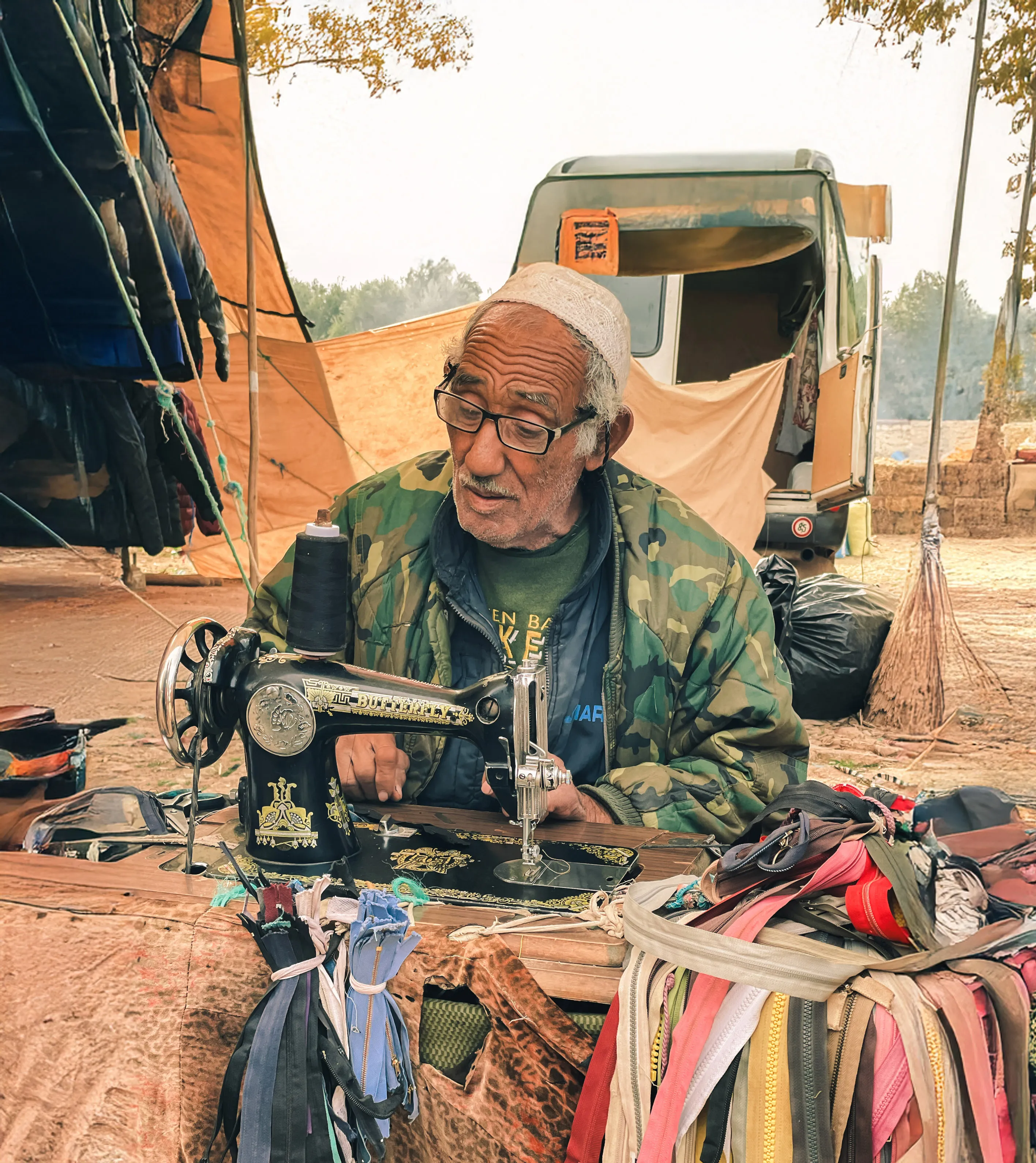 Un homme âgé portant un bonnet et une veste camouflage utilise une machine à coudre ancienne dans un souk en plein air avec des tissus colorés autour au Vallée Ait Bougemez.