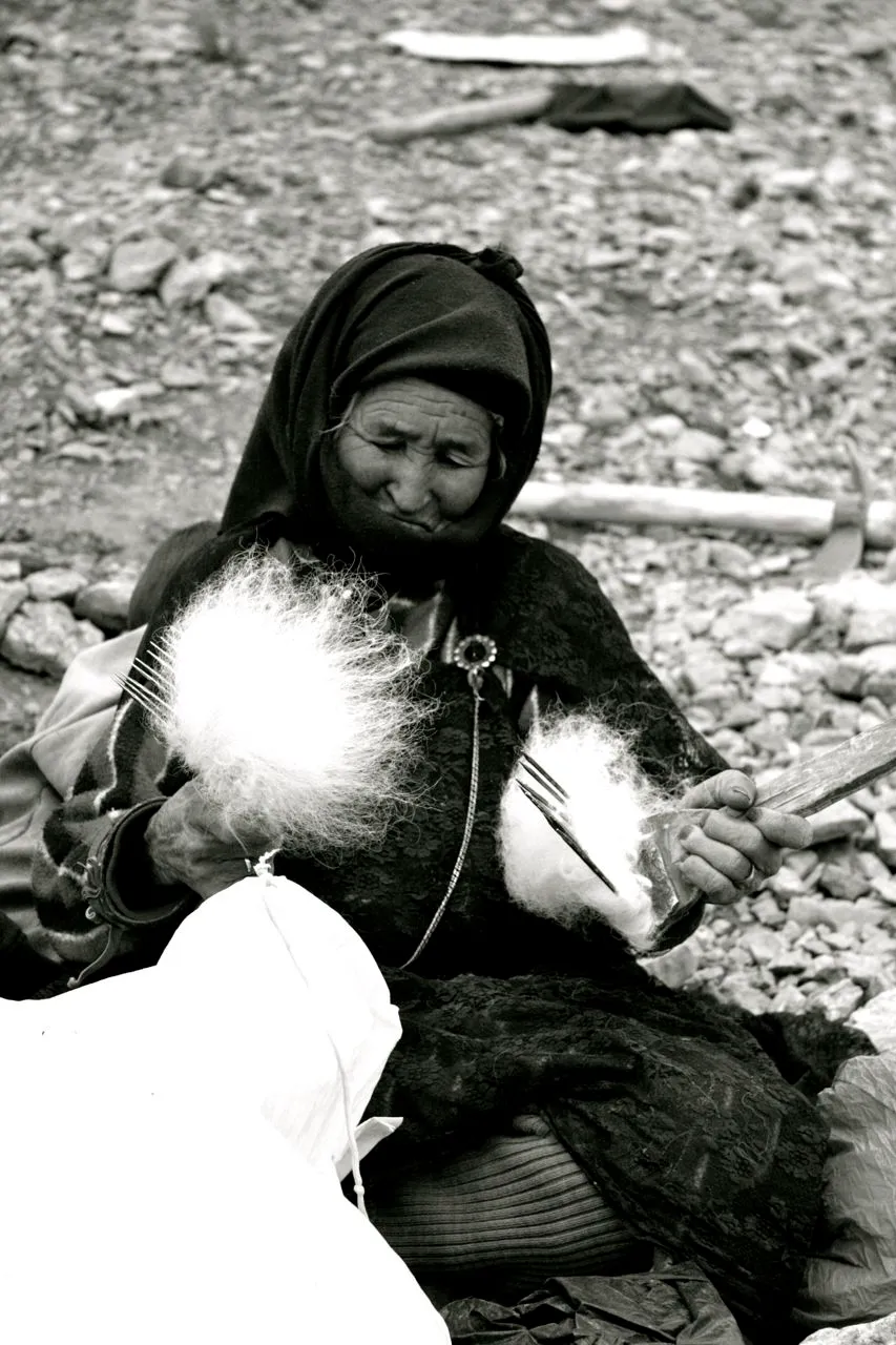 Femme nomade âgée en tête couverte filant de la laine avec des outils traditionnels, assise sur un sol rocheux au haut atlas marocain