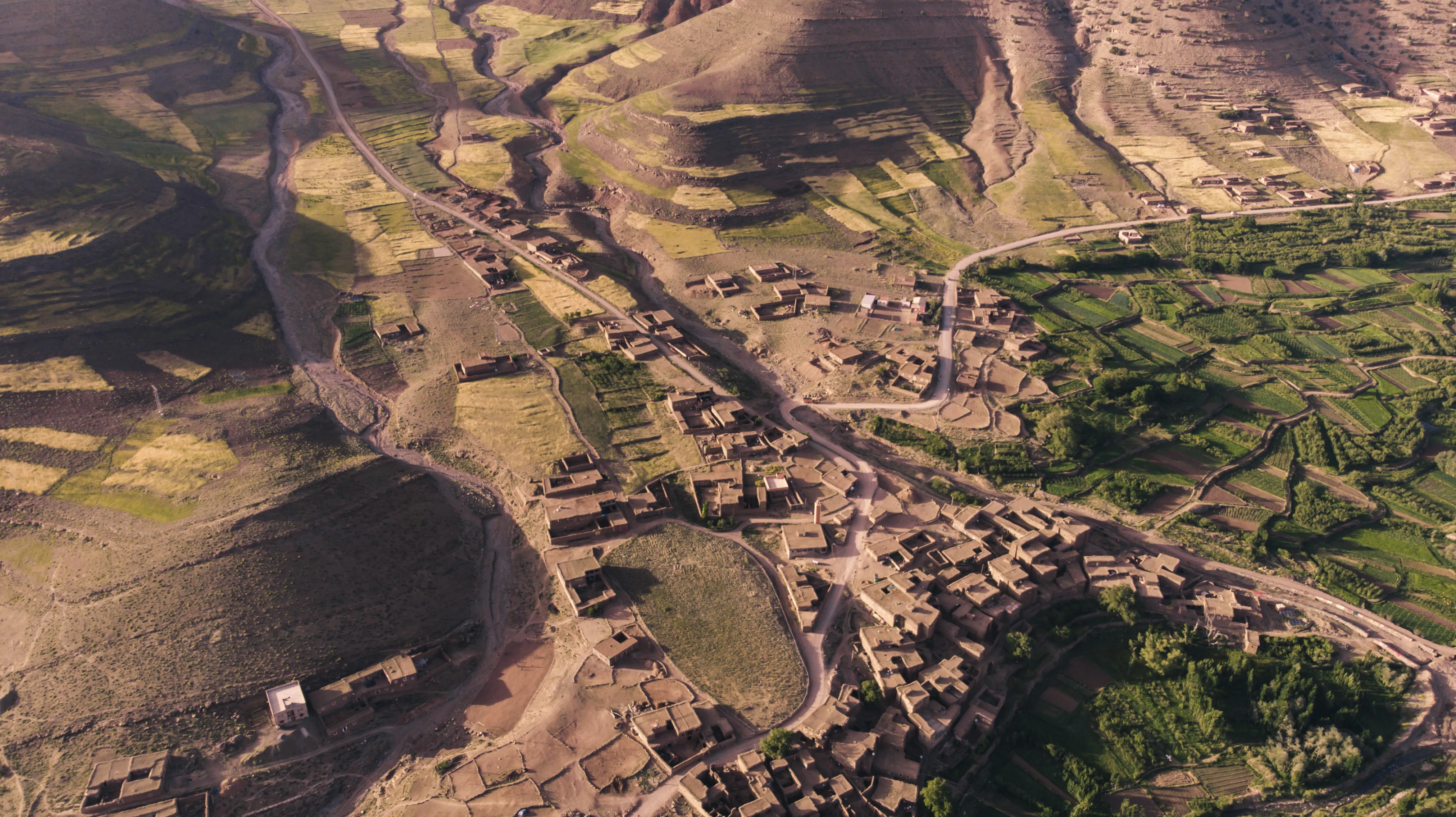 Vue aérienne du village Zawyat Oulmzi dans la vallée ait Bougemez avec des maisons en terre, des routes sinueuses et des champs verts en terrasses.