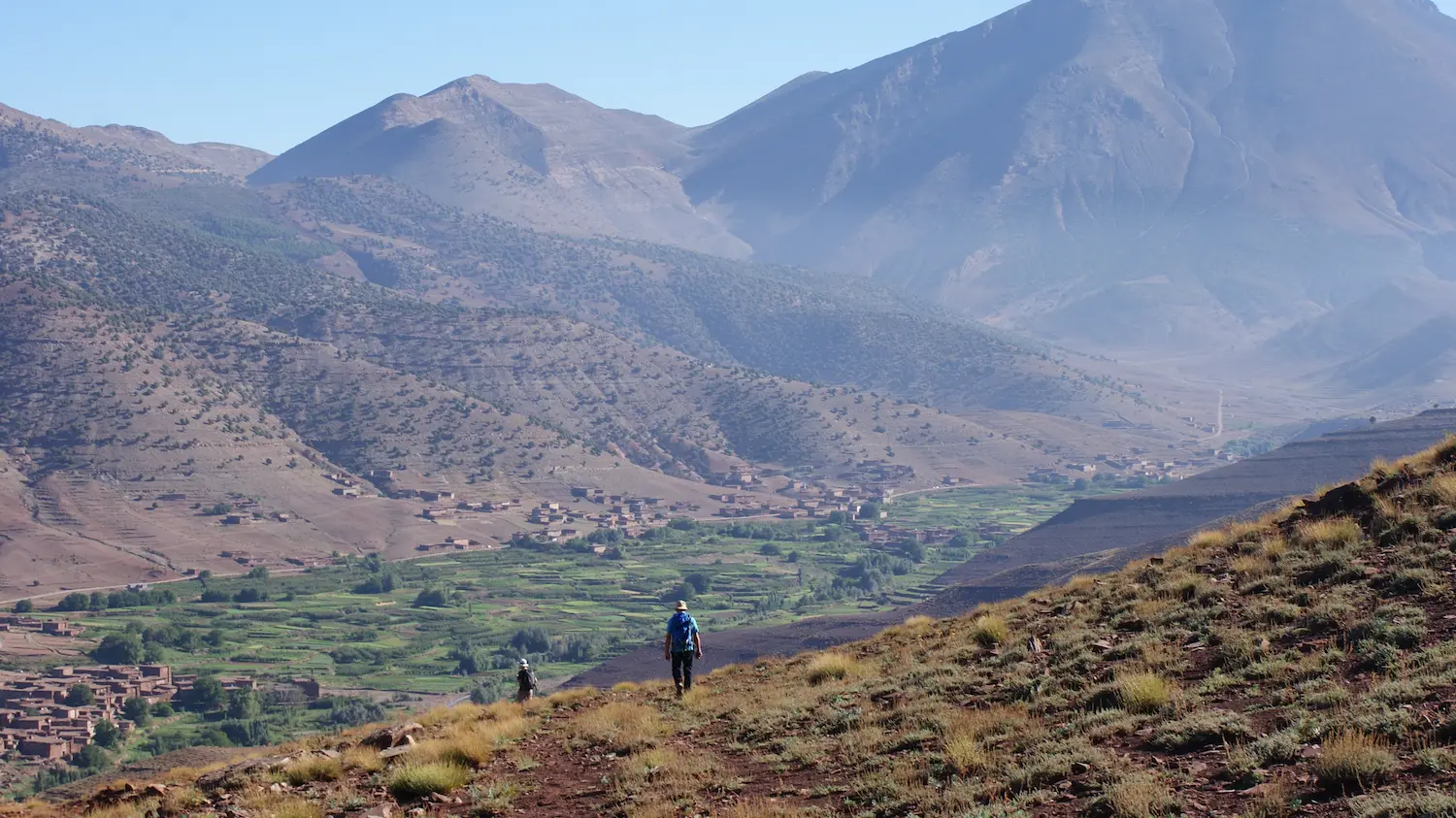 Deux randonneurs marchant sur une colline pierreuse avec un village et des terrasses agricoles dans la vallée Ait Bougemez entourée de montagnes de haut atlas