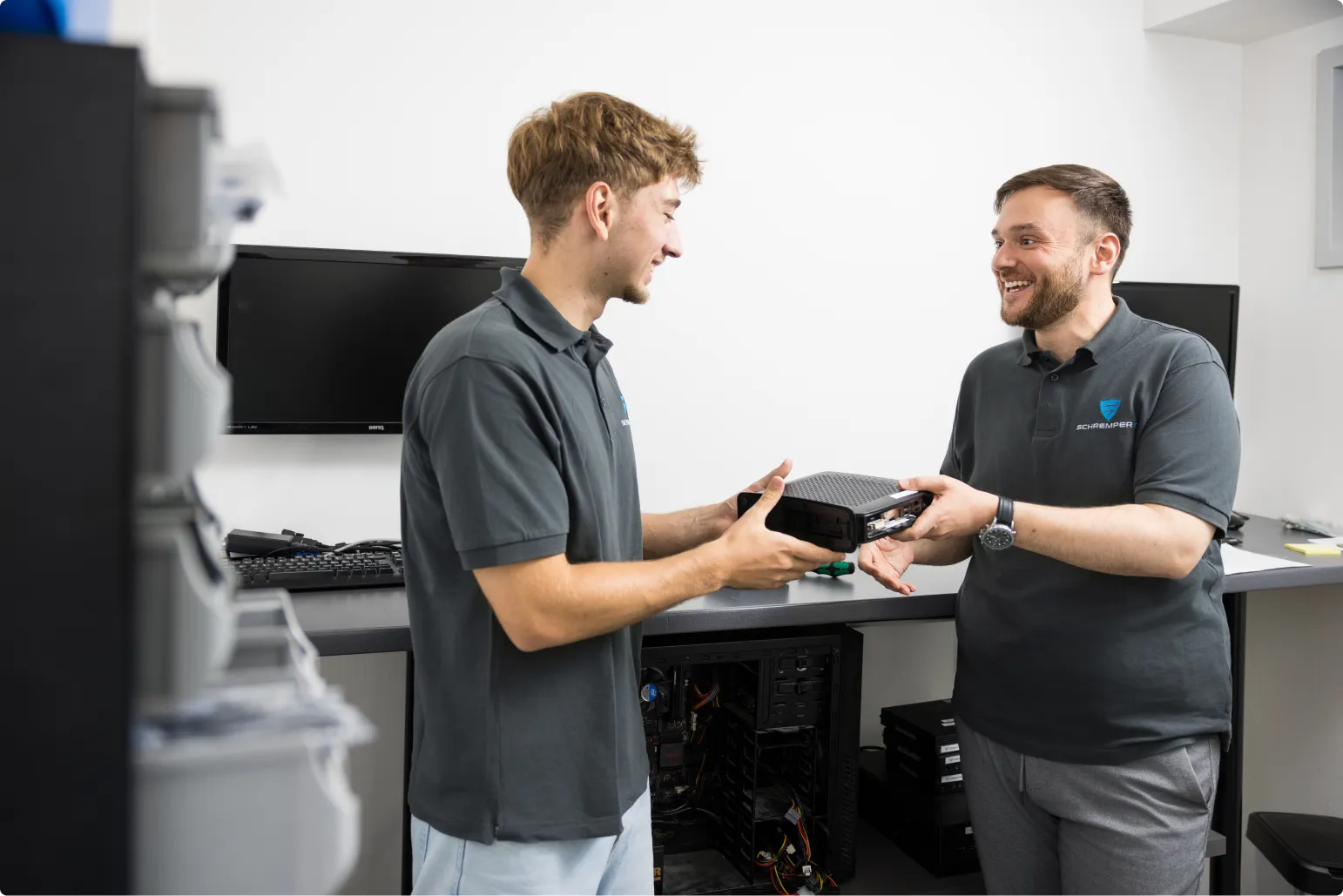 Zwei Mitarbeiter von Schremper IT GmbH stehen in der Werkstatt des IT-Unternehmens in Wuppertal. Ein Mitarbeiter übergibt dem anderen Mitarbeiter die fertig konfigurierte Hardware.