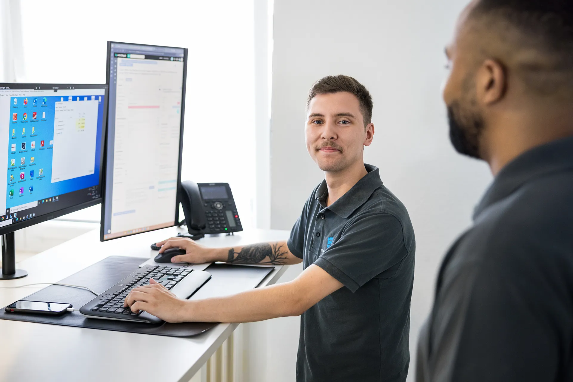 Zwei Mitarbeiter der Schremper IT GmbH stehen vor einem Schreibtisch mit einem klassischen IT-Setup und besprechen ein aktuelles IT-Projekt.