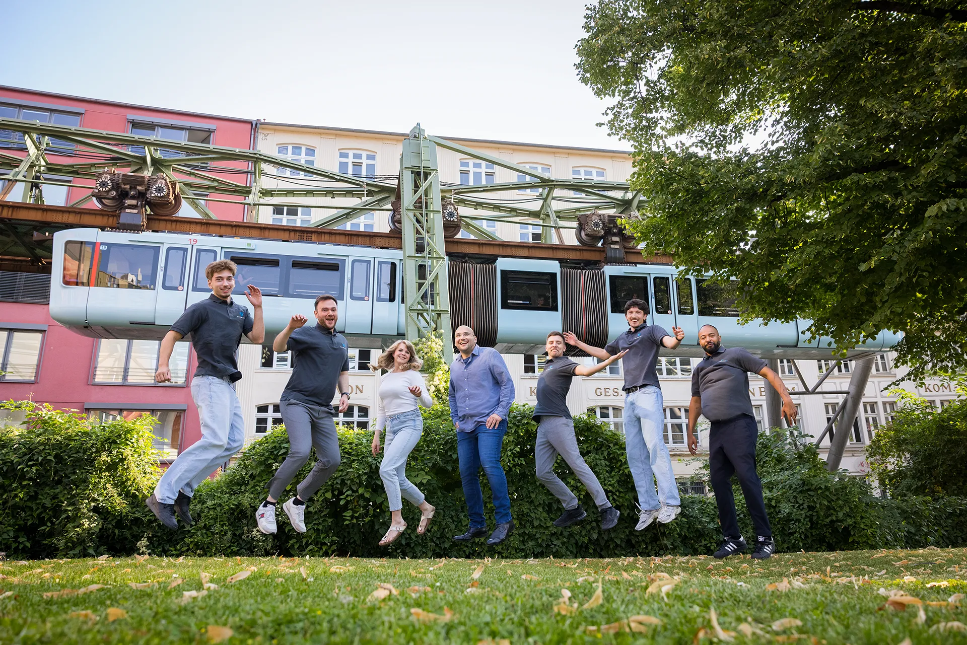 Teamfoto von Schremper IT GmbH in Wuppertal für die Karriereseite. Das Team springt auf einer Rasenfläche vor der Wuppertaler Schwebebahn.