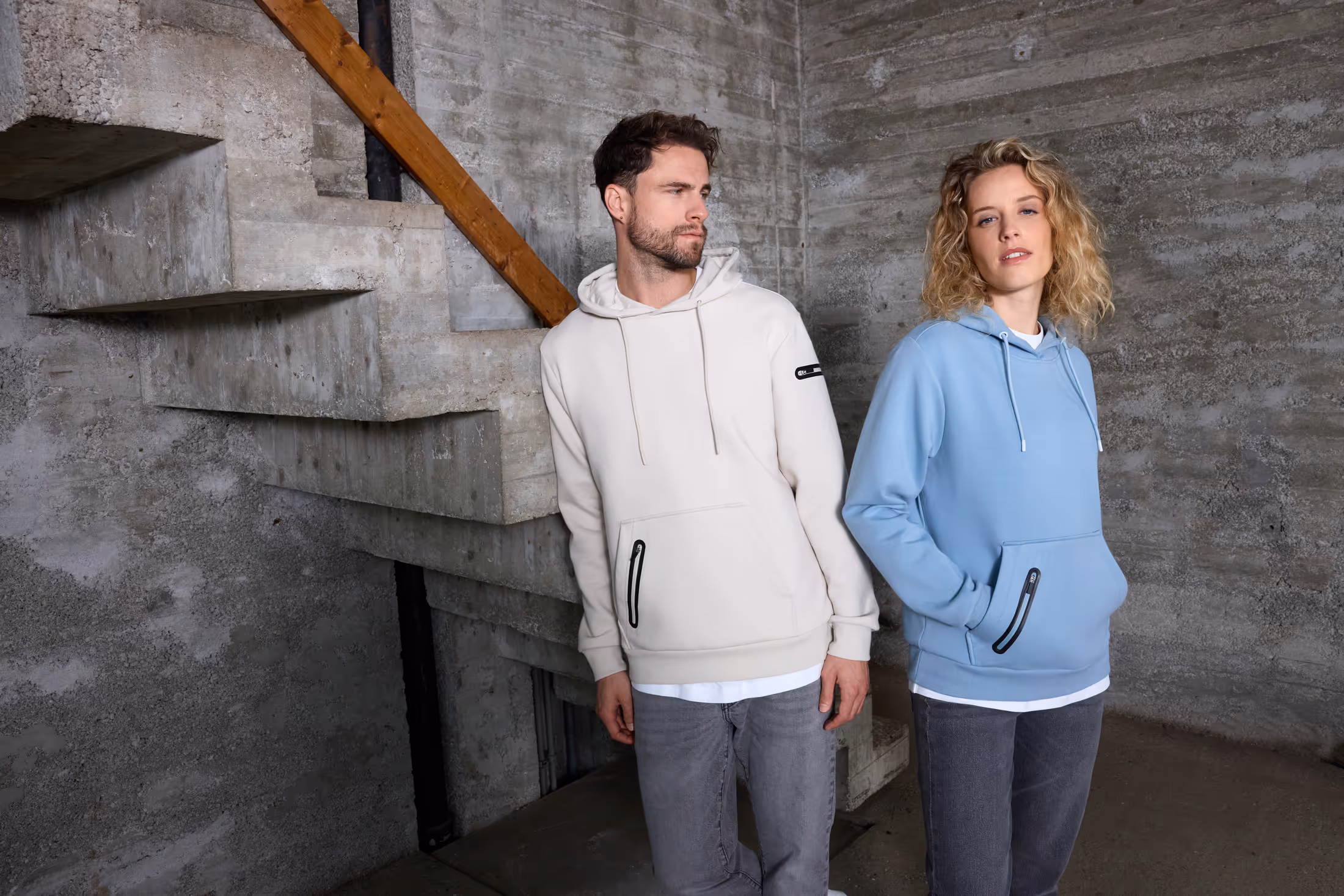 Ein Mann und eine Frau tragen lässige Kapuzenpullover, der Mann in Creme und die Frau in Hellblau, vor einer rohbetonwand mit Treppe.