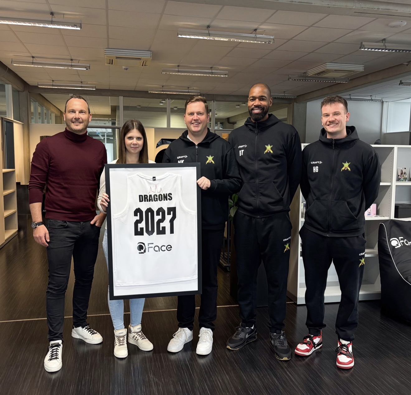 Fünf Personen stehen in einem Büro, zwei in Trainingsanzügen mit Drachen-Logo, eine Frau hält einen eingerahmten weißen Trikot mit der Aufschrift 'DRAGONS 2027 QFace'.