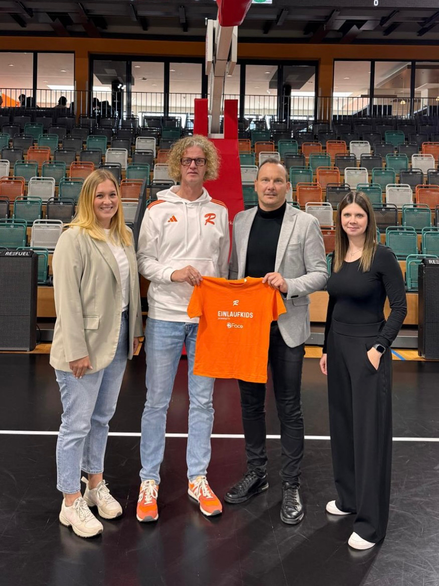 Vier Personen stehen in einer Sporthalle vor leeren Tribünen, zwei halten zusammen ein orangefarbenes T-Shirt mit der Aufschrift 'EINLAUFKIDS'.