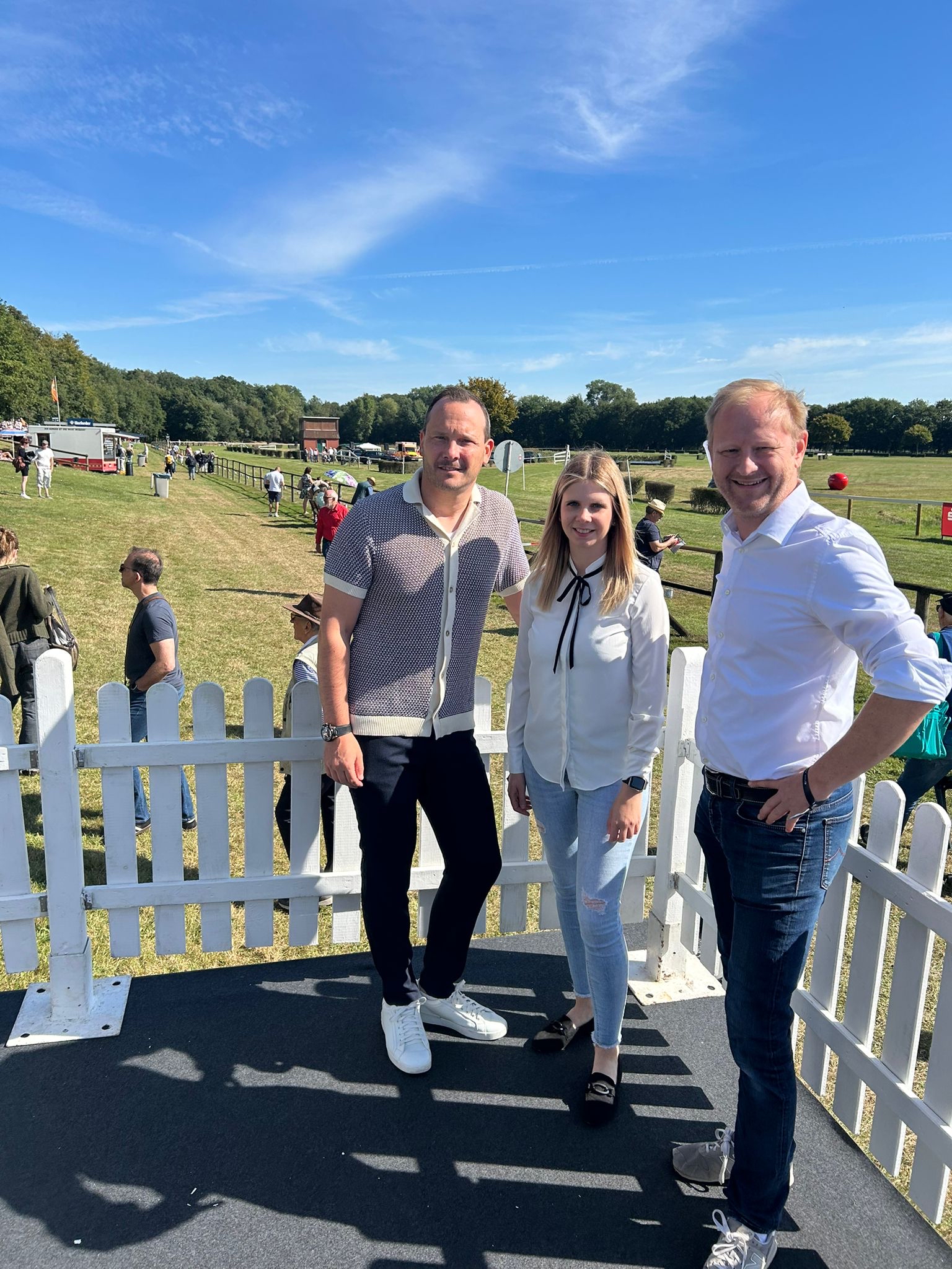 Drei Personen stehen vor einem weißen Lattenzaun auf einer Wiese bei sonnigem Himmel.