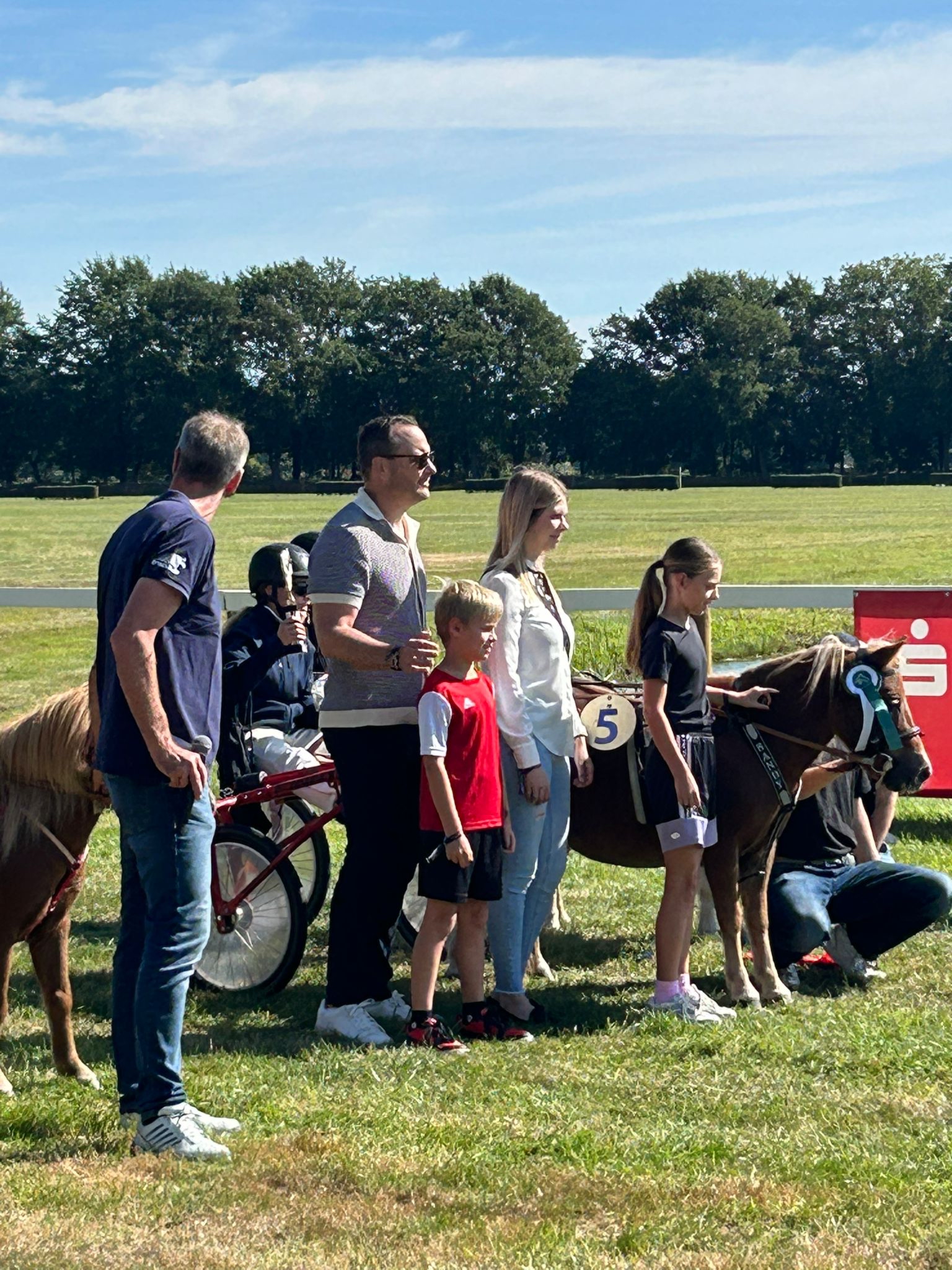 Eine Gruppe von Menschen steht auf einer Grasfläche neben zwei Ponys mit Startnummern, bei sonnigem Wetter.