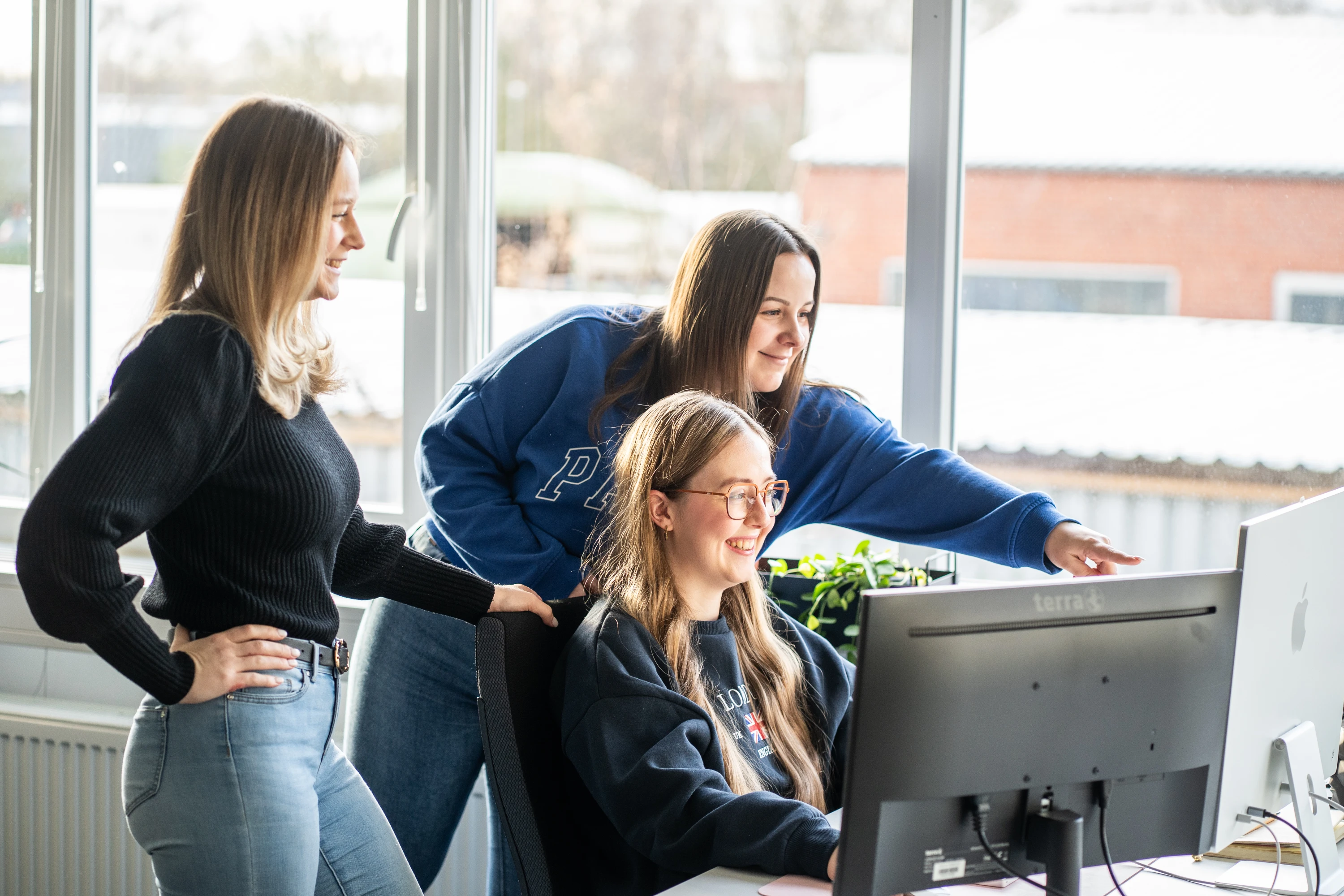 Drei junge Frauen arbeiten gemeinsam an einem Computer in einem hellen Büro mit großen Fenstern.
