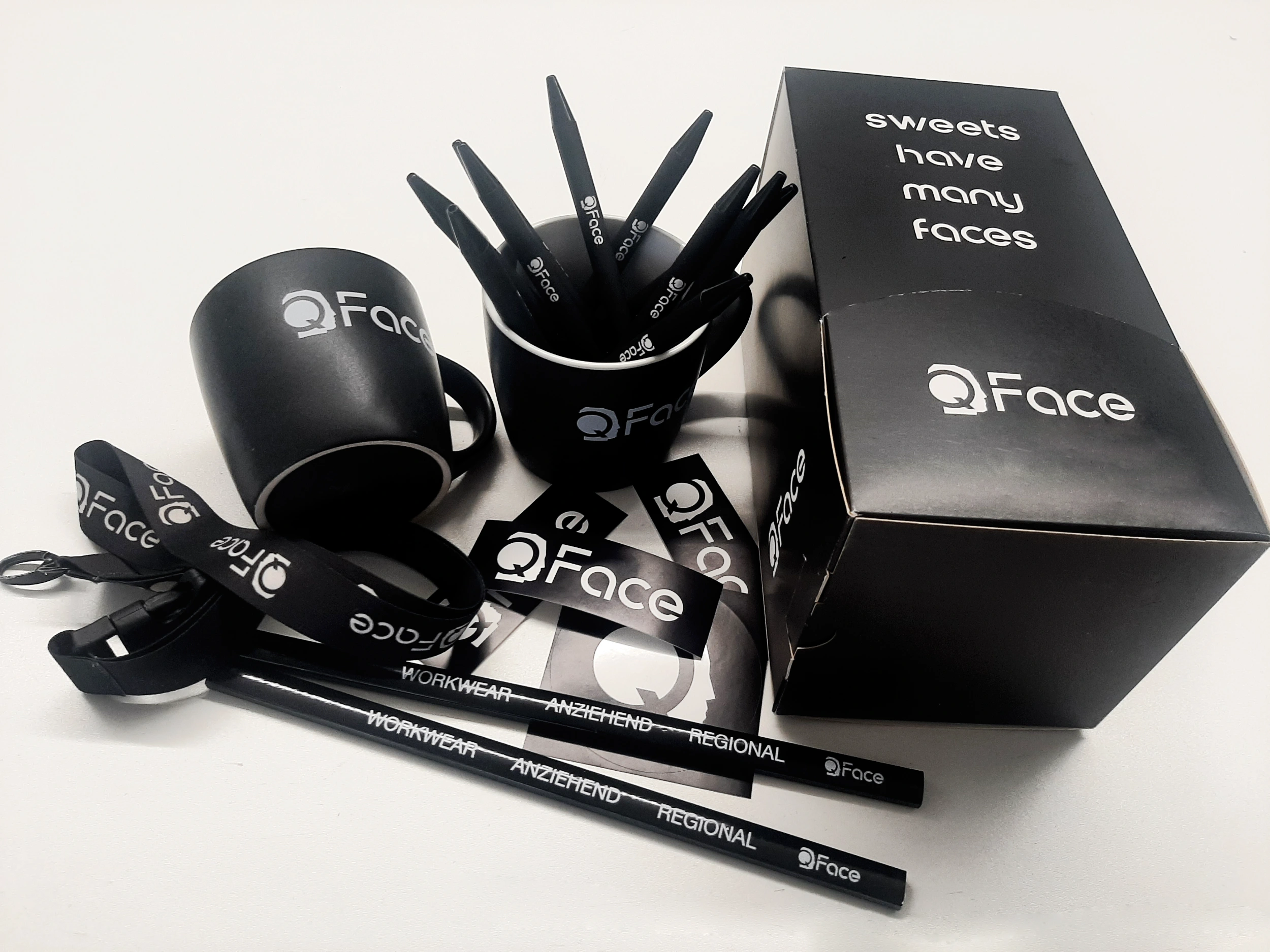 Schwarze Werbeartikel mit dem QFace-Logo, darunter Tassen, Stifte, Schlüsselbänder und eine Box mit dem Schriftzug ‚sweets have many faces‘.