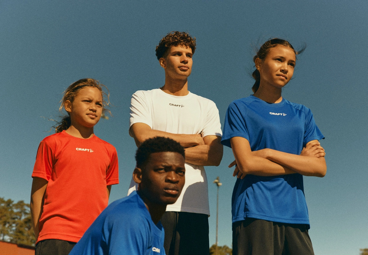 Vier Jugendliche in verschiedenen Sportswear-T-Shirts stehen und sitzen draußen bei klarem Himmel.