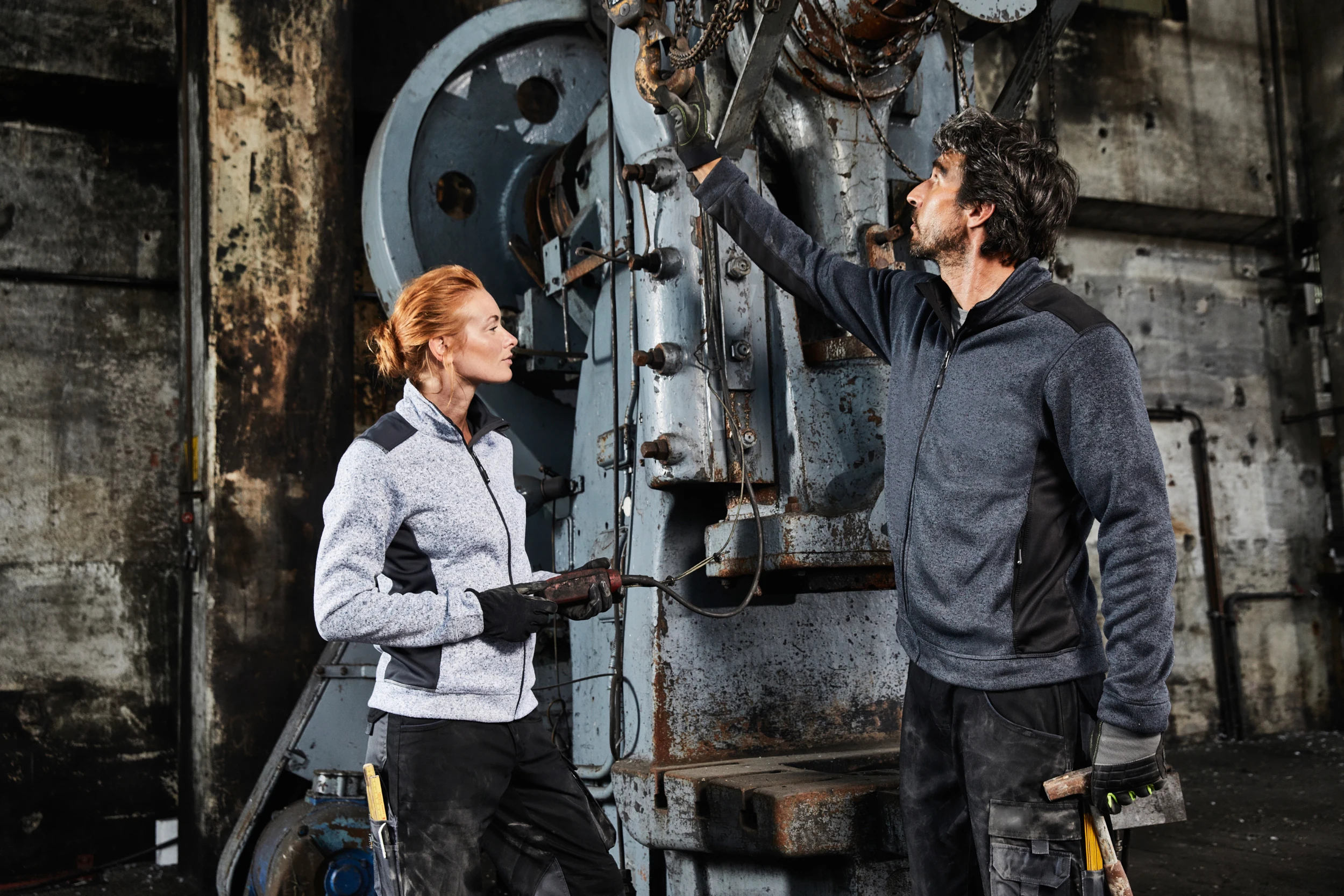 Zwei Industriearbeiter in Arbeitskleidung inspizieren und bedienen eine große, alte Maschine in einer Fabrikhalle.