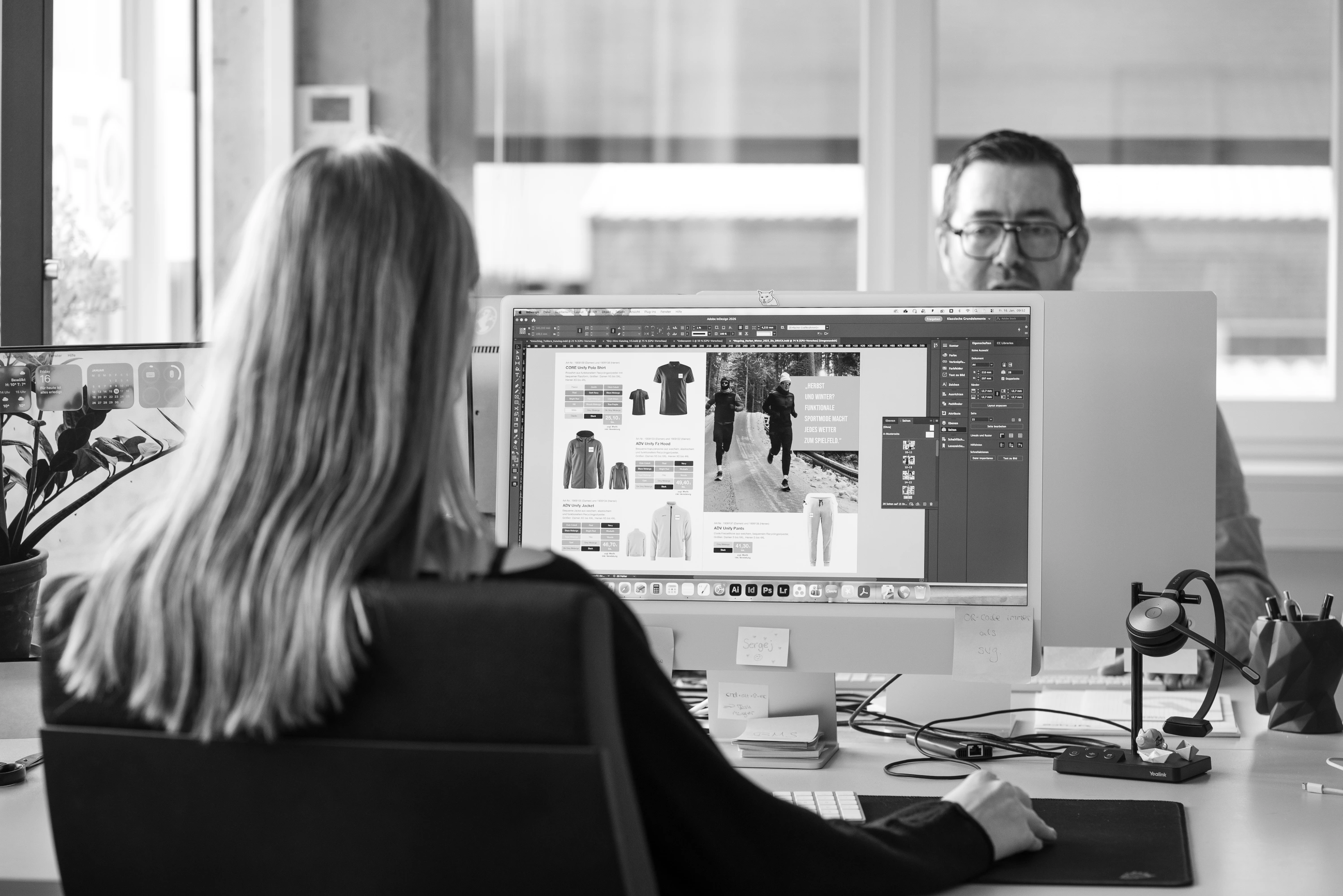 Frau bearbeitet am Computer ein Design mit Bekleidungsprodukten, Mann sitzt ihr gegenüber an einem Büroarbeitsplatz.