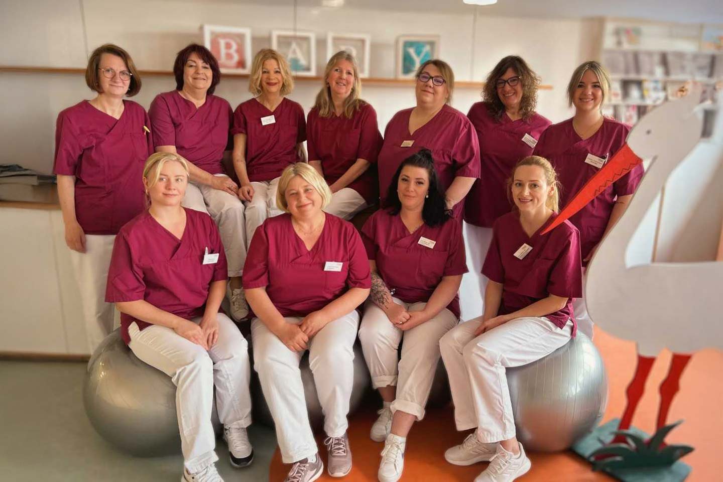 Elf Frauen in roten Pflegeuniformen und weißen Hosen, einige sitzen auf Gymnastikbällen, Gruppenfoto in einem hellen Raum mit einem Storch aus Holz.