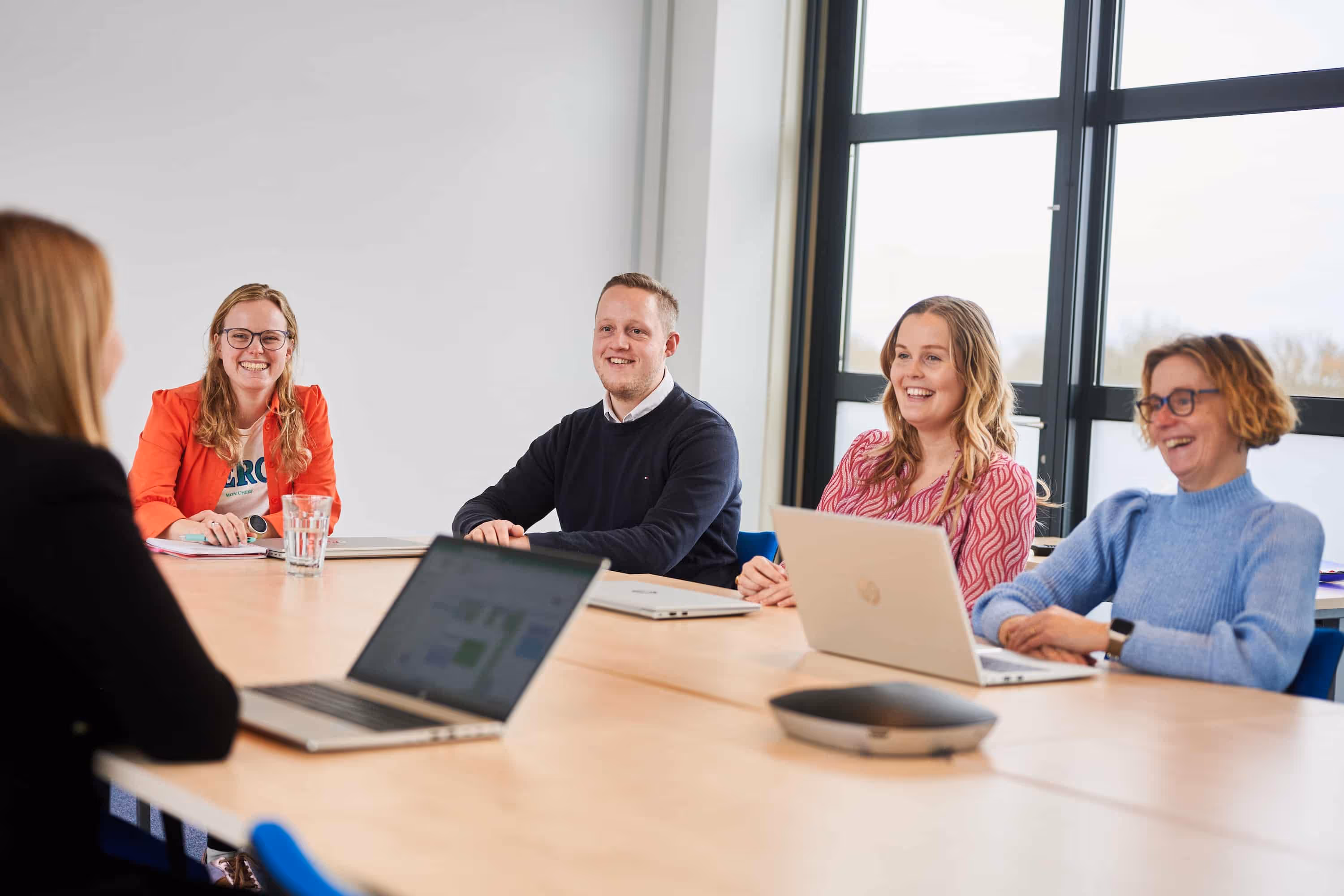 Vier lachende volwassenen zitten aan een vergadertafel met laptops en notitieblokken in een lichte kantoorruimte.